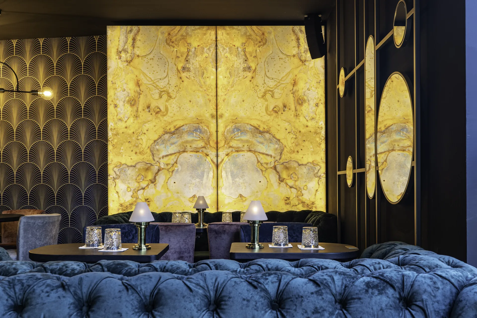 Sitzbereich der Nuba Bar in Linz mit blauem Chesterfield-Samtsofa, gedeckten Tischen mit goldenen Tischlämpchen und einem imposanten hinterleuchteten gelben Marmorpaneel vor Art-Déco-Tapete.