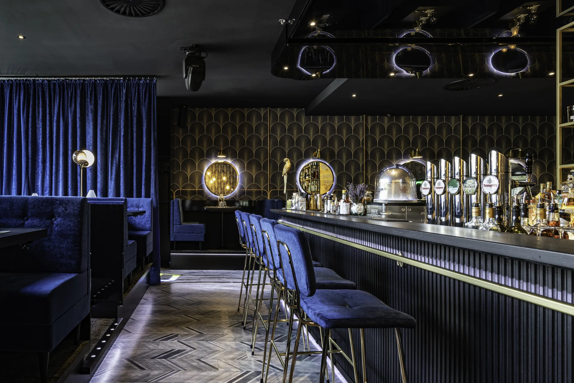Blick entlang der Nuba Bar in Linz mit blauen Samtbarhockern, Zapfanlage, beleuchteten Rundspiegeln und Art-Déco-Goldtapete vor blauem Samtvorhang auf dem charakteristischen Fischgrätboden.