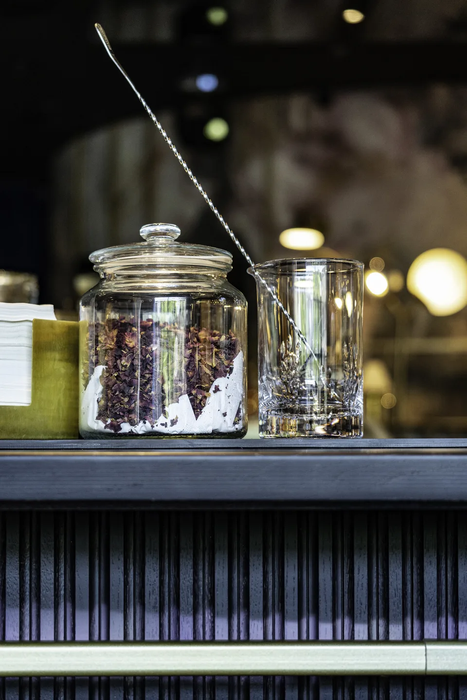 Detailaufnahme auf dem Bartresen der Nuba Bar in Linz mit einem Glasgefäß voller getrockneter Rosenblütenblätter und einem Mixglas mit Barlöffel.