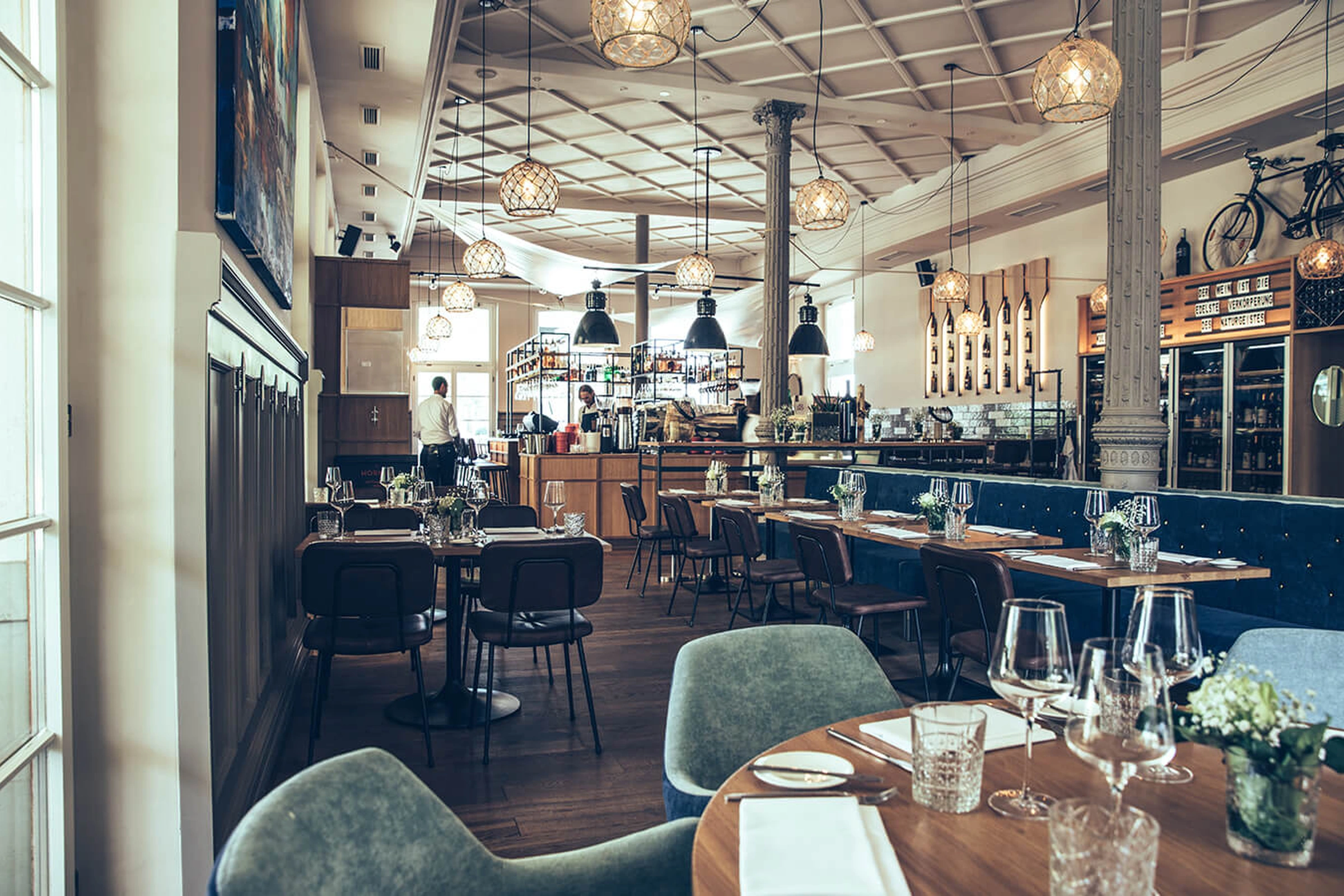 Gesamtansicht des Restaurant Pier 69 in Bregenz mit gedeckten Holztischen, mintgrünen Sesseln, blauer Sitzbank und Korbpendelleuchten in einem großzügigen hellen Gastraum.