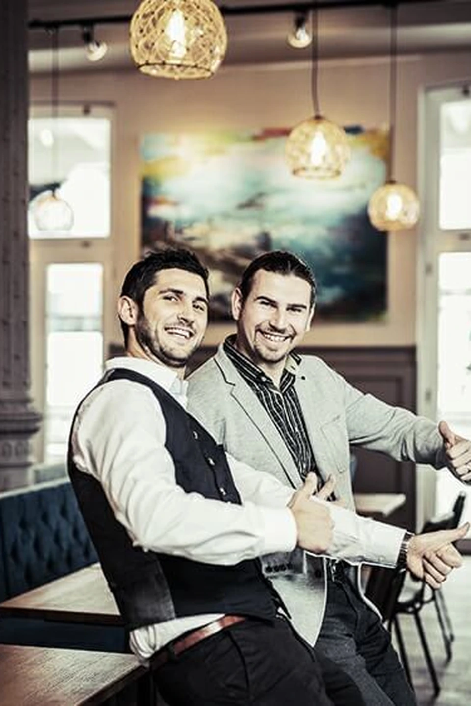 Die zwei Inhaber des Restaurant Pier 69 in Bregenz posieren lächelnd im Gastraum vor einem farbenfrohen Gemälde und den charakteristischen Korbpendelleuchten.