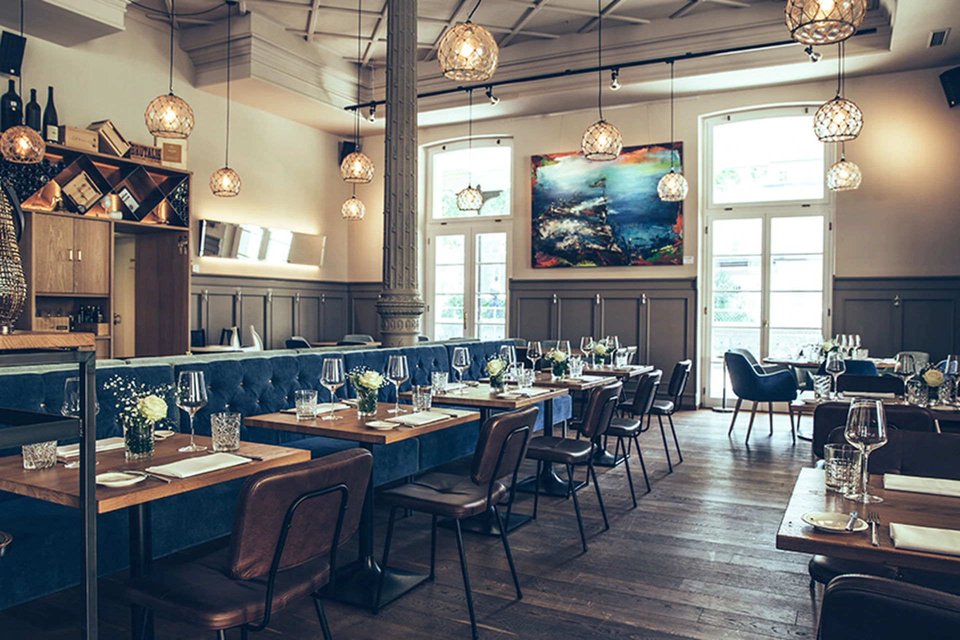 Gastraum des Restaurant Pier 69 in Bregenz mit blauer Chesterfield-Sitzbank, gedeckten Holztischen, braunen Lederstühlen, einem großformatigen Kunstgemälde und Korbpendelleuchten.