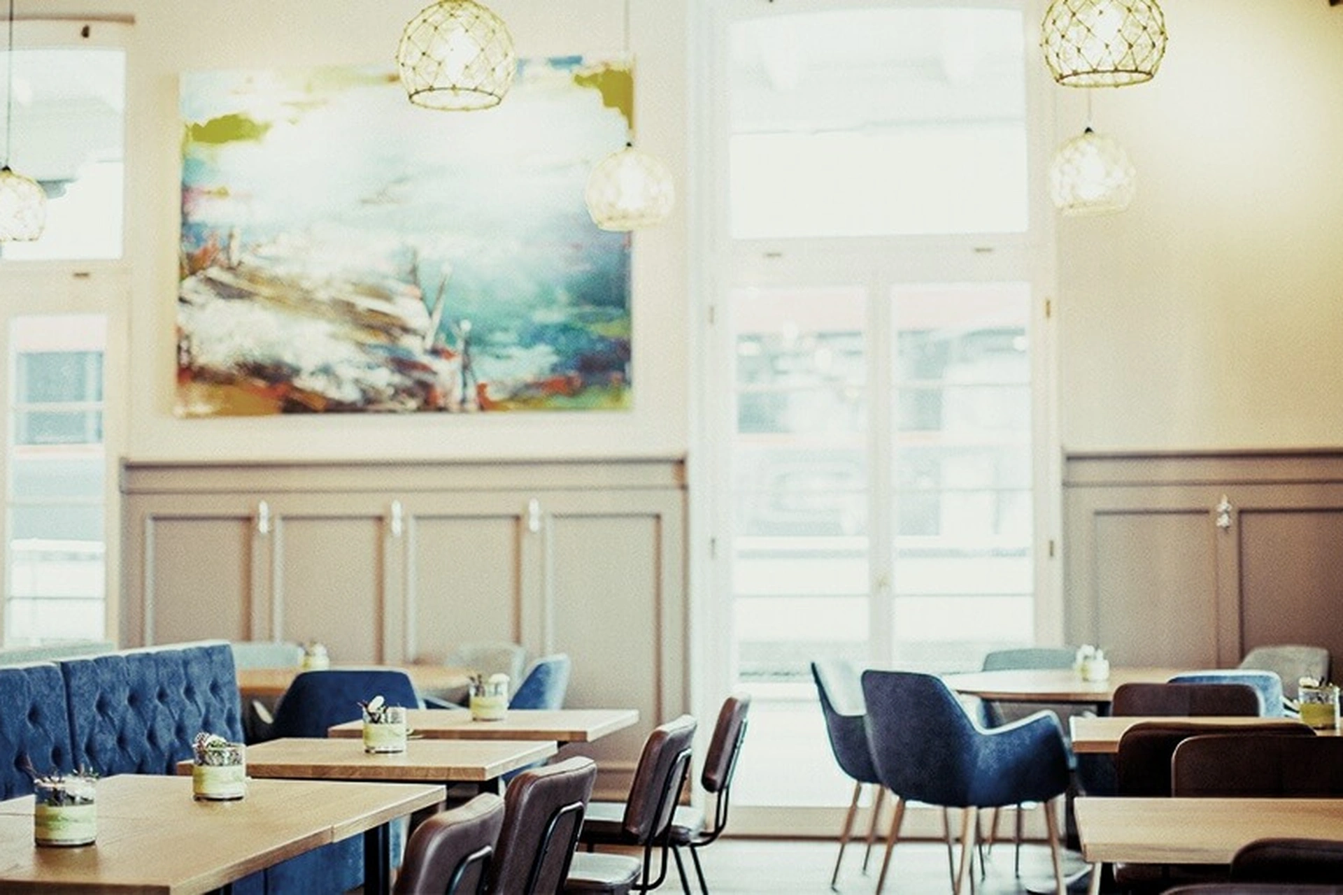Heller Gastraum des Restaurant Pier 69 in Bregenz mit blauer Chesterfield-Sitzbank, gemischten Sesseln, großformatigem Kunstgemälde und Panoramafenstern mit Tageslicht.