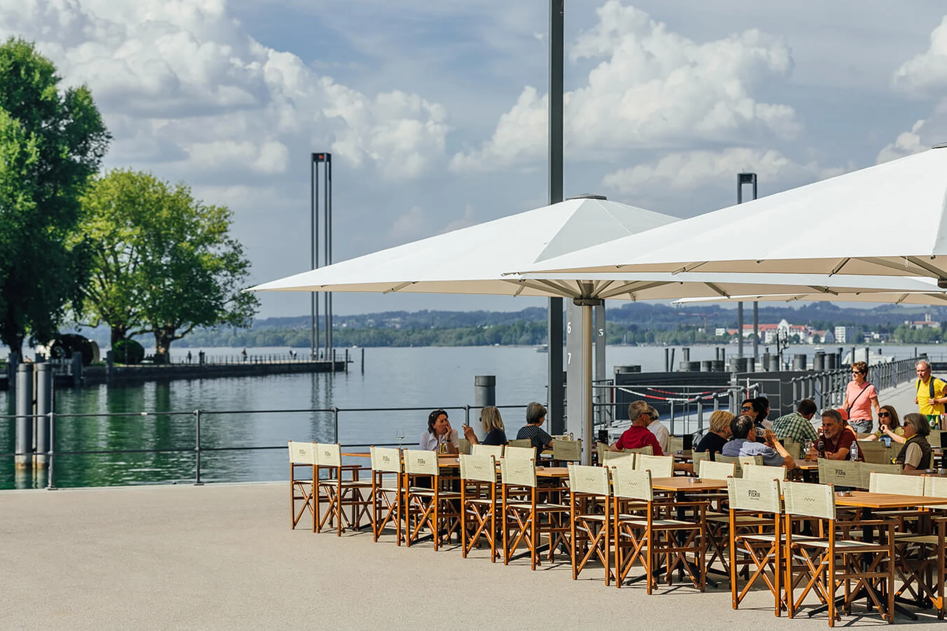 Seeterrasse des Restaurant Pier 69 in Bregenz mit großen weißen Sonnenschirmen, Holz-Regiesesseln und Gästen mit Blick auf den Bodensee.