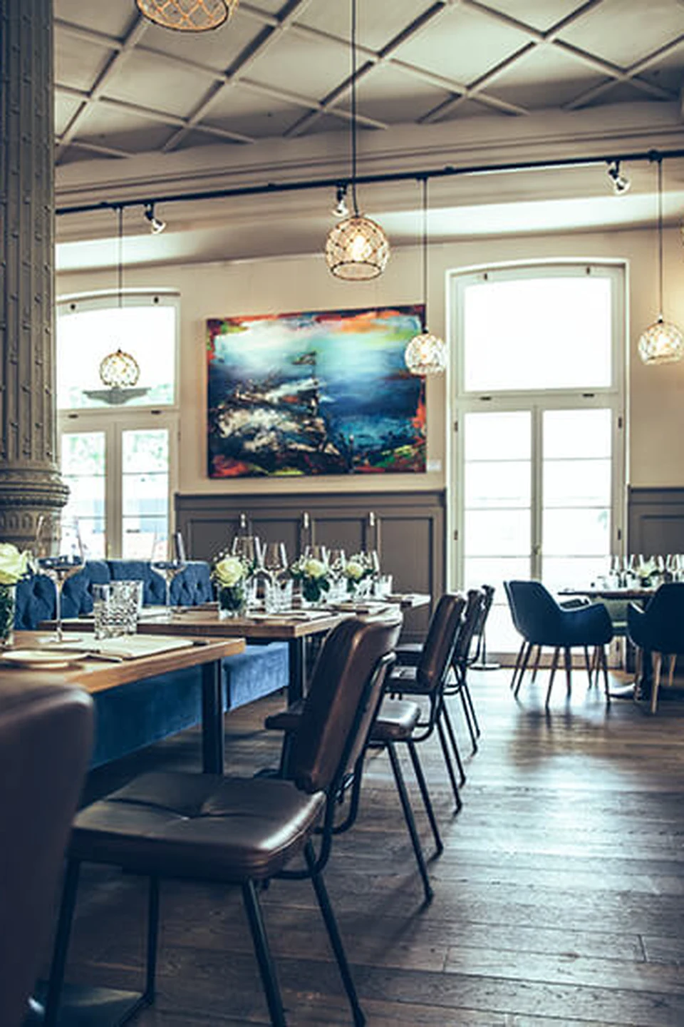Gastraum des Restaurant Pier 69 in Bregenz mit blauer Chesterfield-Sitzbank, gedeckten Holztischen, braunen Lederstühlen, großformatigem Kunstgemälde und Korbpendelleuchten über dem Altholzboden.