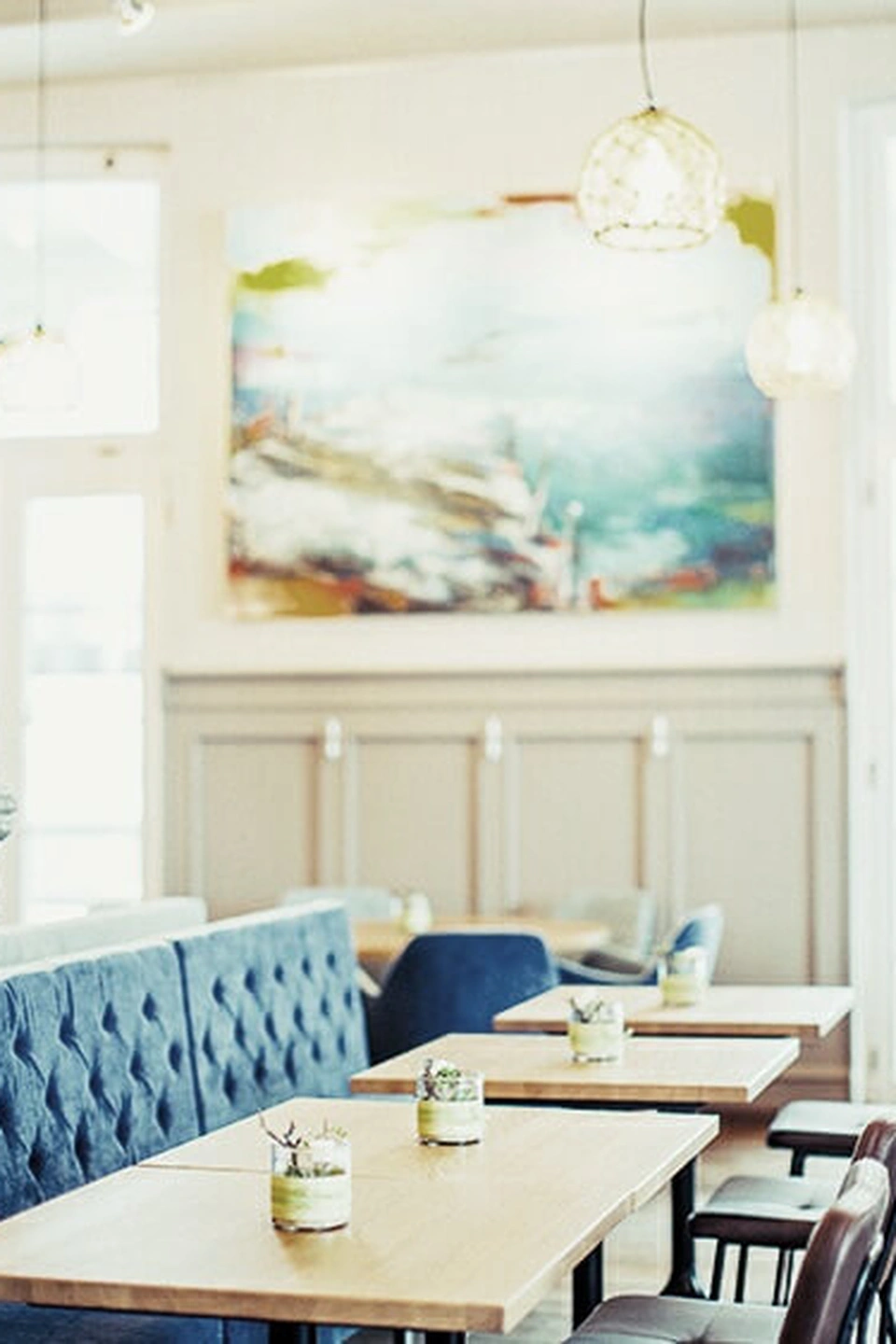 Heller Sitzbereich des Restaurant Pier 69 in Bregenz mit blauer Chesterfield-Sitzbank, hellen Holztischen, einer Korbpendelleuchte und einem farbenfrohen Kunstgemälde an der Wand.