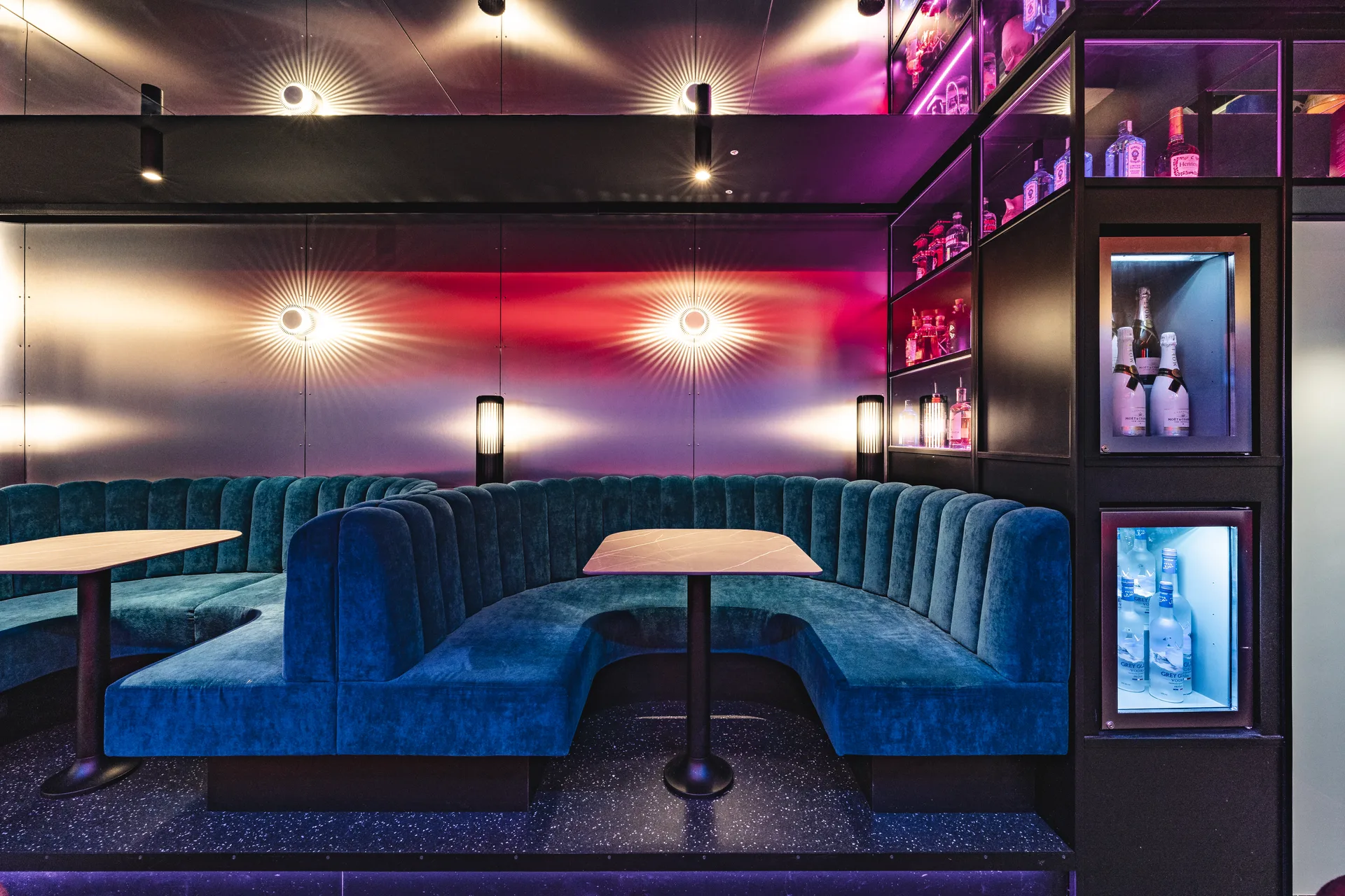 Loungebereich des Club Pure in Kufstein mit blauer geschwungener Samtbank, runden Wandleuchten mit Strahlenkranz-Effekt und beleuchtetem Spirituosenregal in pinkfarbenem Ambientelicht.