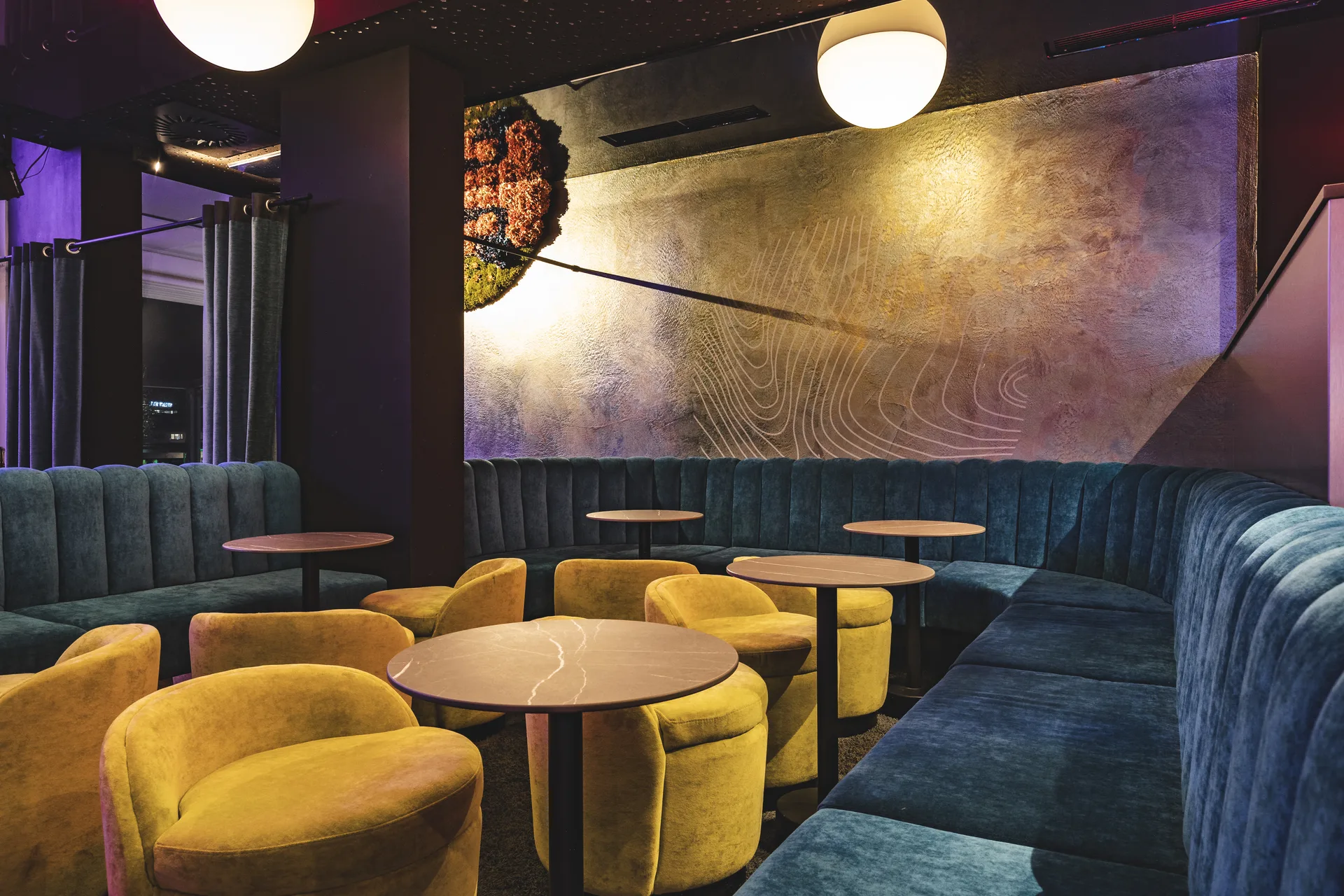 Loungebereich des Club Pure in Kufstein mit gelben Samtsesseln, blauer Kanalbank, marmorieren Rundtischen und einem großflächigen Wandmotiv mit Jahresringen-Grafik unter schwebenden Moosbild-Installationen.