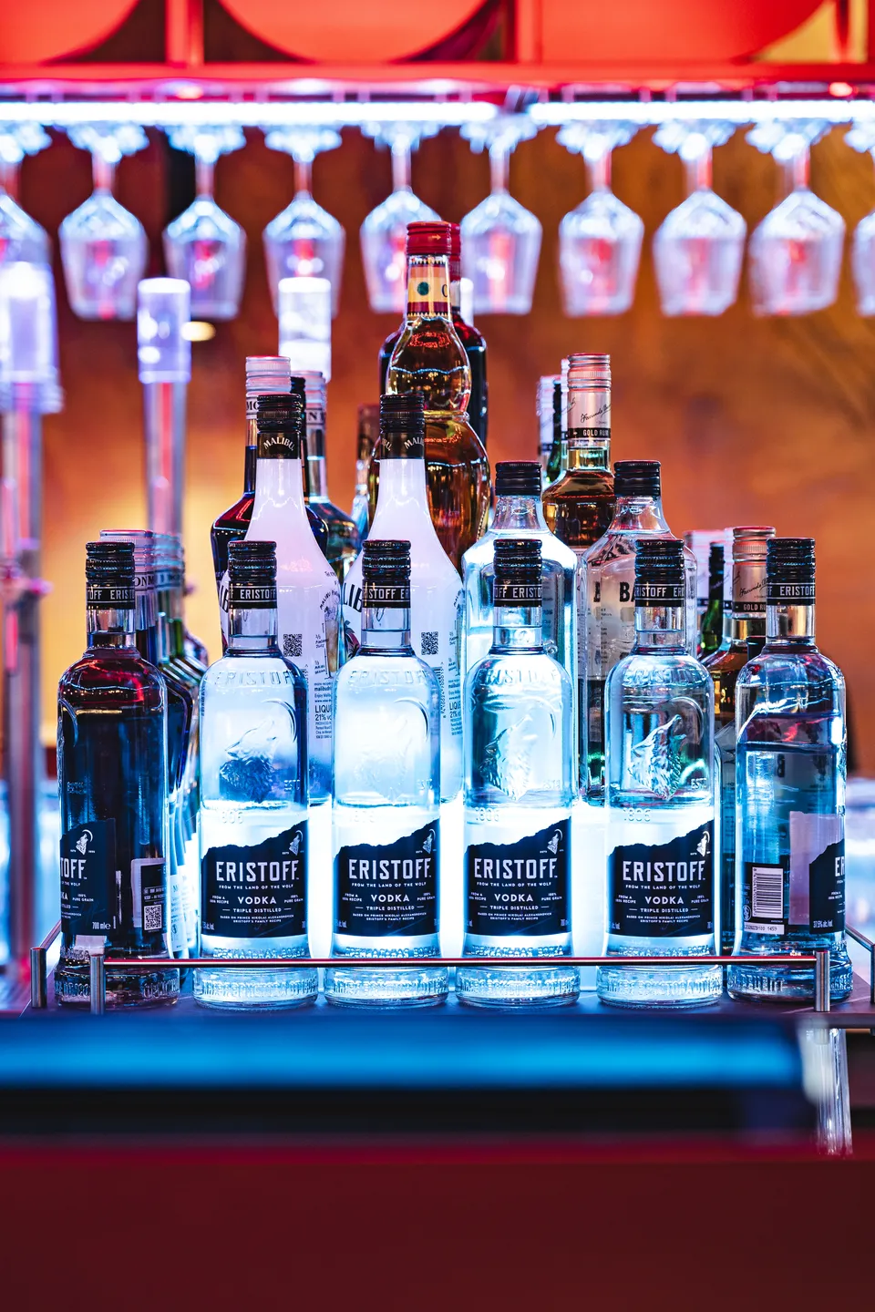 Beleuchtetes Spirituosenregal des Club Pure in Kufstein mit mehreren Eristoff Vodka-Flaschen in blauem Licht, darüber hängende Weingläser vor roter Deckeninstallation.