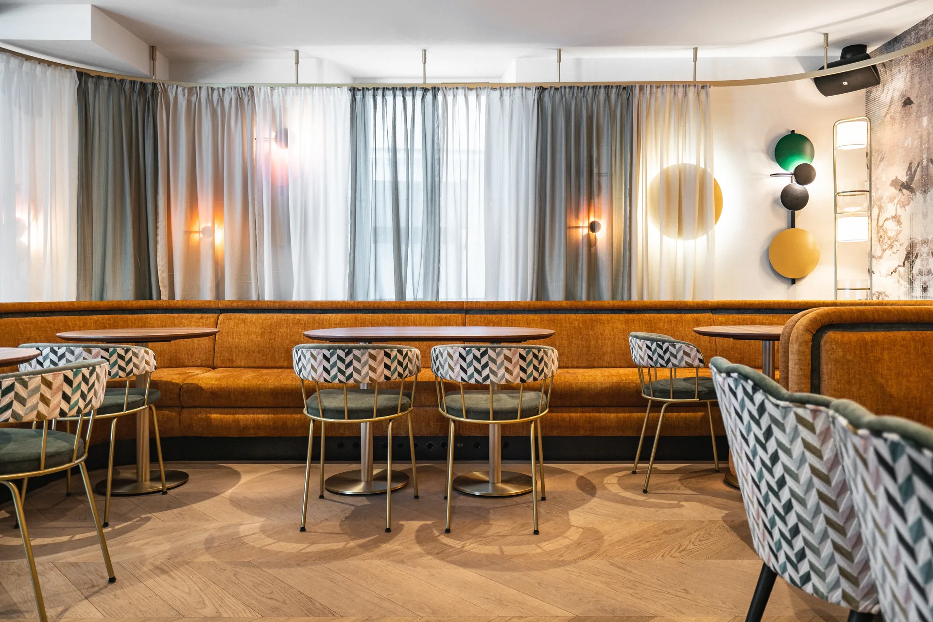 Gastraum der Bar Rio in Dornbirn mit durchgehender orangefarbener Samtbank, gemusterten Goldrahmen-Sesseln, lichten Vorhängen und einer verspielten Wandleuchten-Komposition aus farbigen Scheiben.