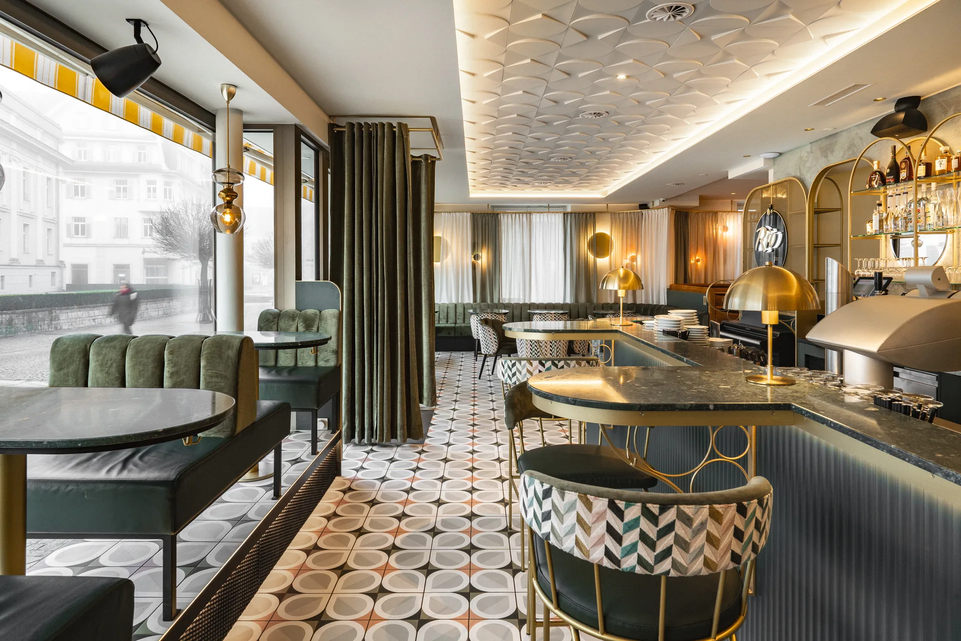 Gesamtblick in die Bar Rio in Dornbirn mit grünem Marmortresen, gemusterten Barhockern, grüner Samtbank, Panoramafenster zur Straße und farbenfrohem Zementfliesenboden unter der strukturierten Decke.