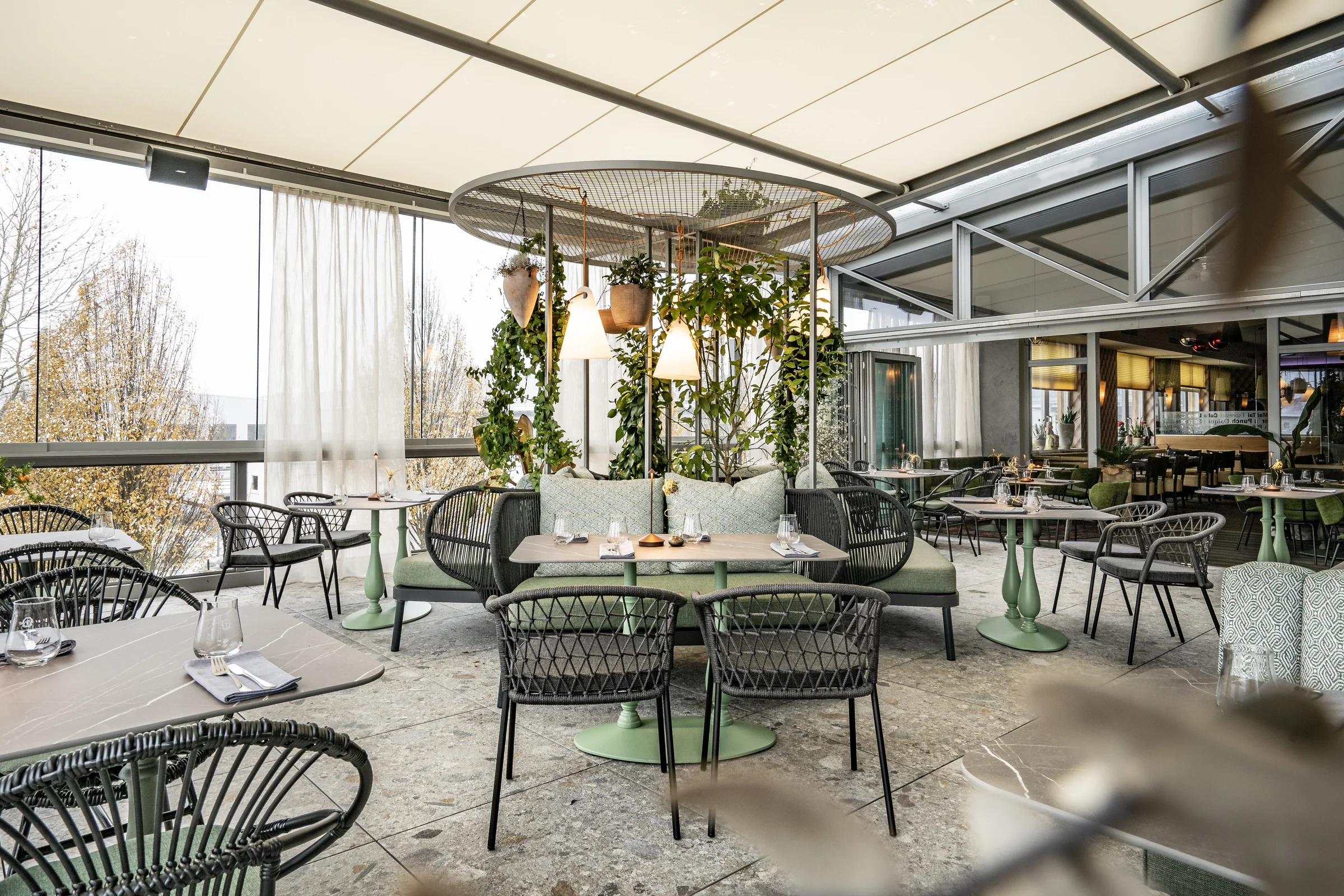 Überdachter Wintergarten des Restaurant Sam's in Eggelsberg mit geflochtenen Korbsesseln, mintgrünen Tischbeinen, üppigen Hängepflanzen und Panoramafenstern mit Blick auf den herbstlichen Außenbereich.