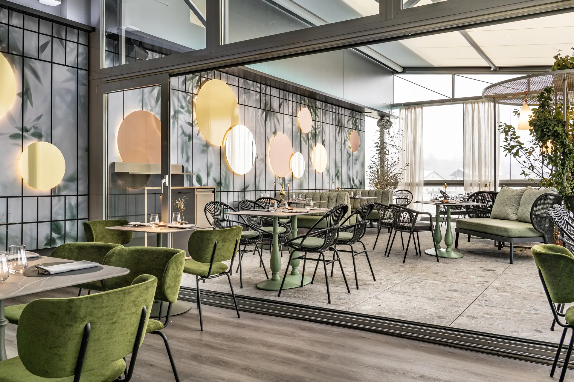 Weitläufiger Gastraum des Restaurant Sam's in Eggelsberg mit grünen Samtsesseln und Korbsesseln, runden Wandspiegeln, Botanik-Tapete und großen Panoramafenstern.