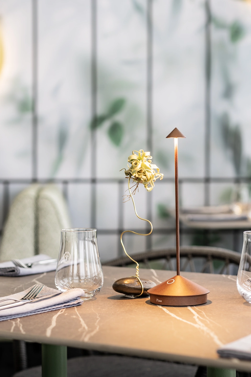 Detailaufnahme eines gedeckten Tisches im Restaurant Sam's in Eggelsberg mit Weingläsern, Leinenservietten, kupferfarbener Tischlampe und Marmorplatte vor Botanik-Tapete.