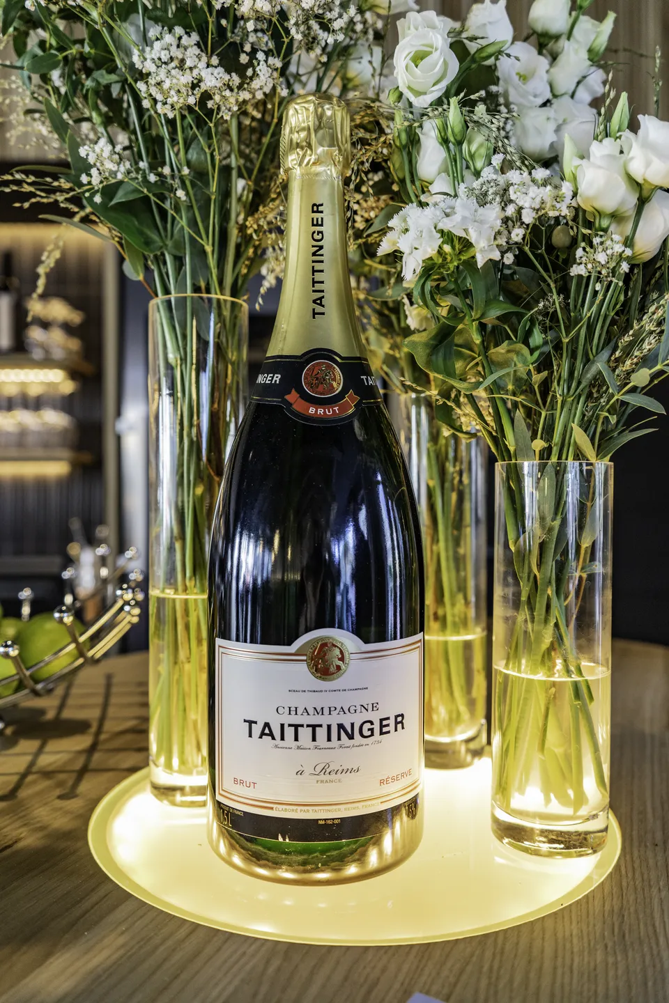 Detailaufnahme einer Taittinger Champagnerflasche zwischen beleuchteten Glasvasen mit weißen Blumen auf dem Bartresen des Scanaua.