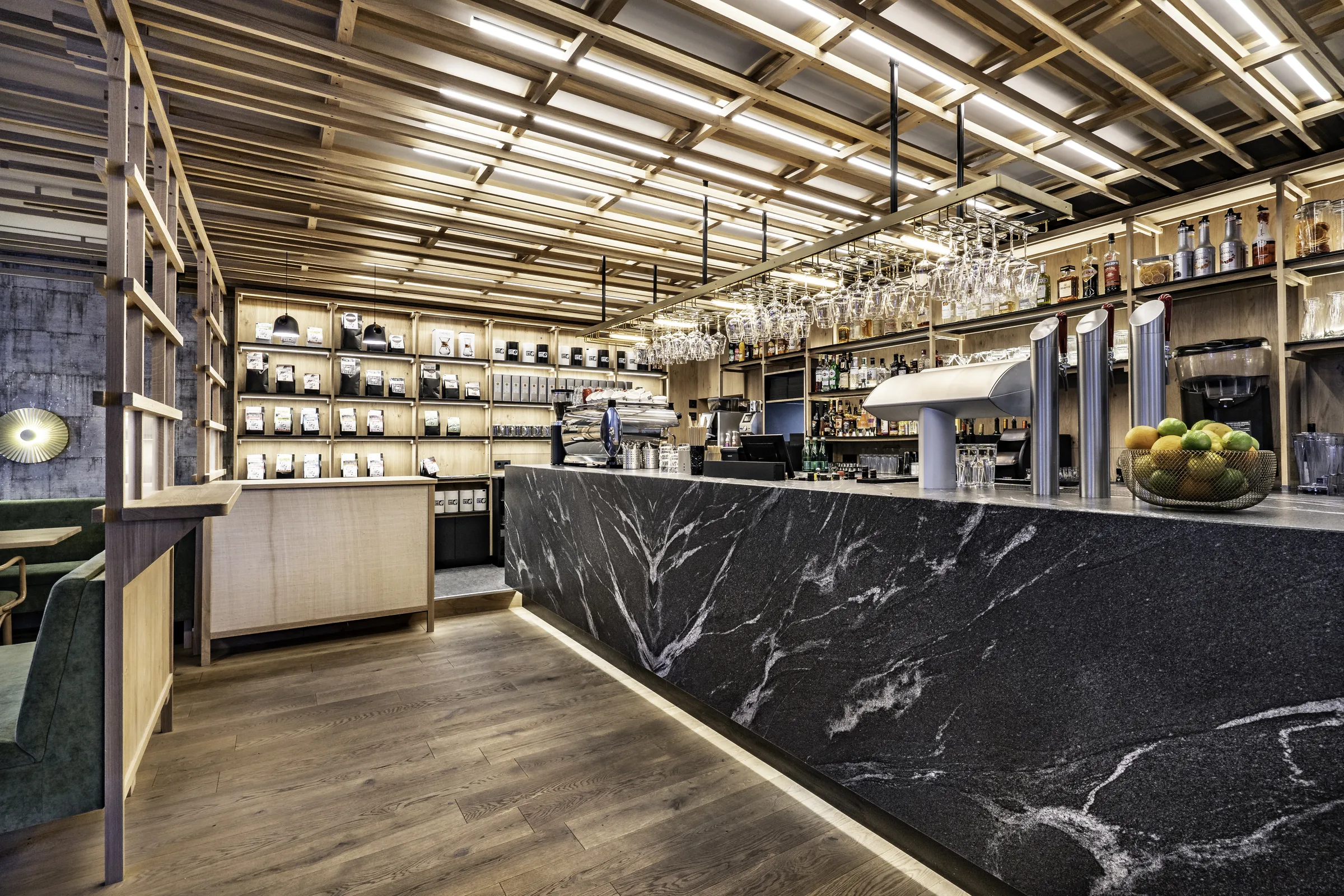 Bar des Café 21 in Dornbirn mit schwarzem Marmortresen, hängenden Weingläsern, beleuchtetem Holzregal mit Kaffeeprodukten und Espressomaschine.