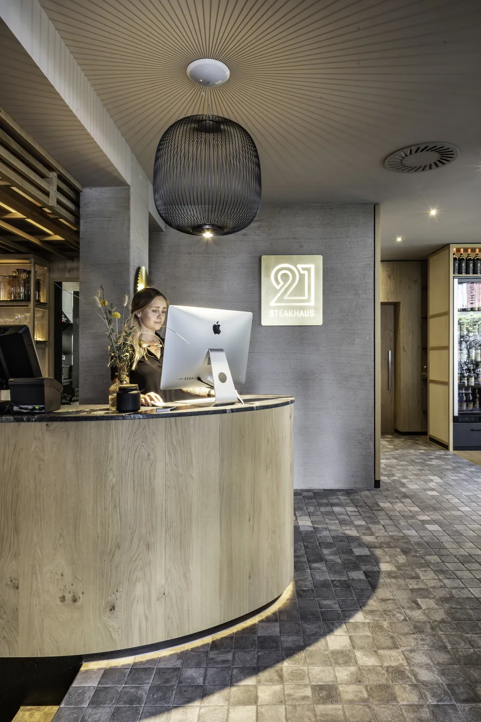 Empfangsbereich des Steakhaus 21 in Dornbirn mit rundem Holztresen, Mitarbeiterin am iMac, leuchtendem „21 Steakhaus