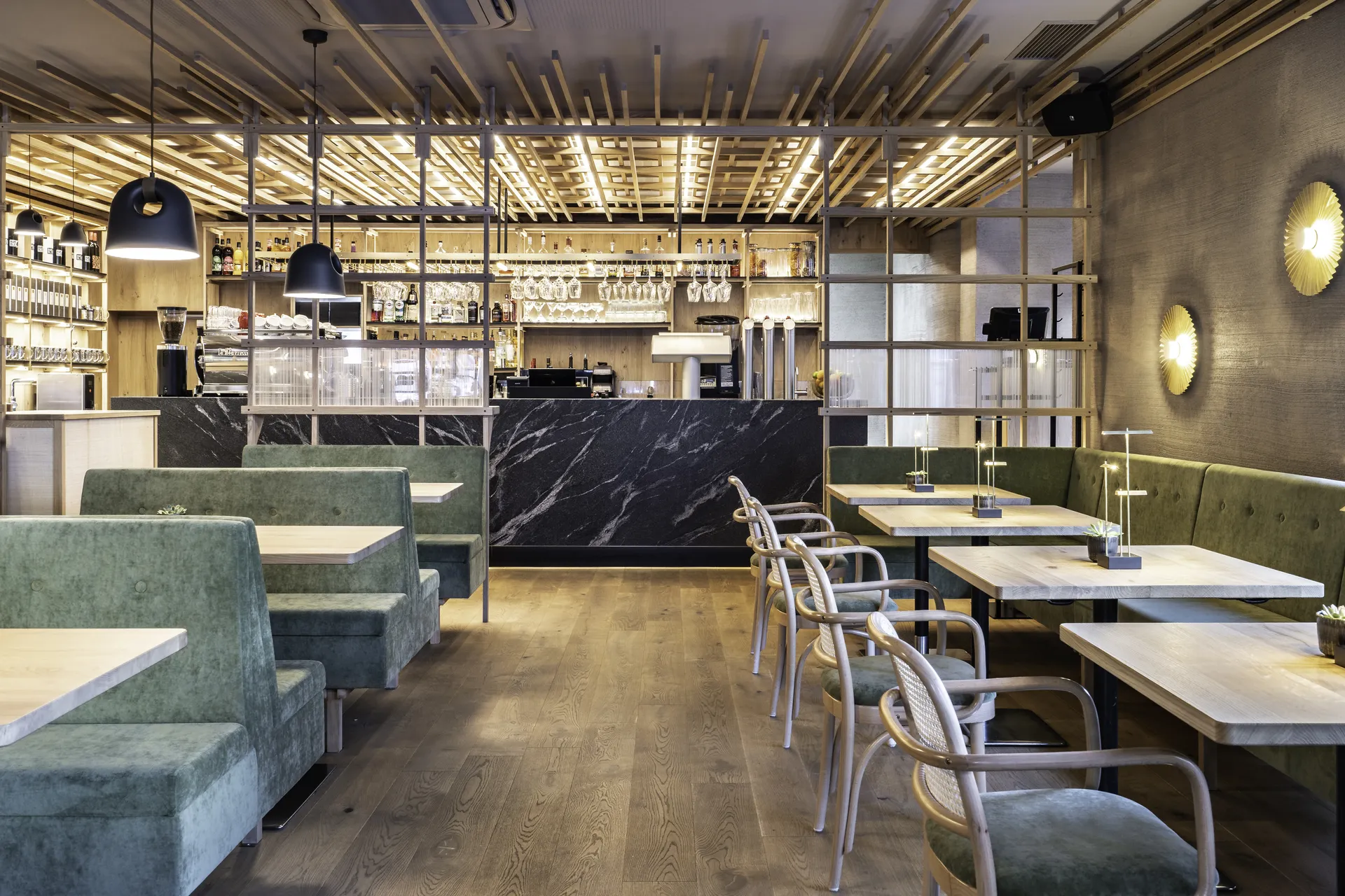 Gastraum des Café 21 in Dornbirn mit grünen Samtbänken, schwarzem Marmortresen, hängenden Weingläsern und beleuchtetem Holzlatten-Deckenraster.