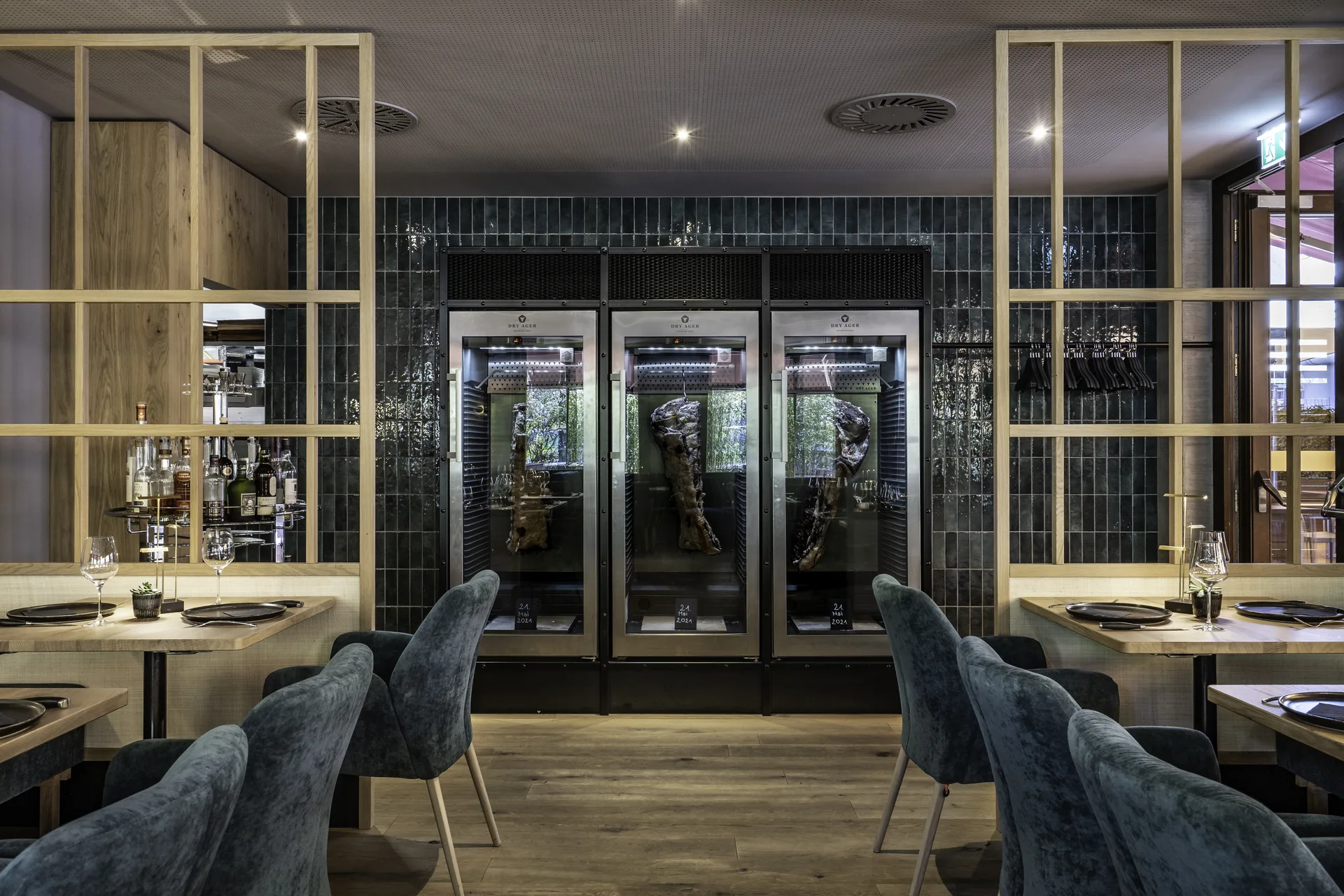 Gastraum des Steakhaus 21 in Dornbirn mit drei Dry-Ager-Reifeschränken, blauen Samtsesseln, gedeckten Holztischen und dunkler Fliesenrückwand.