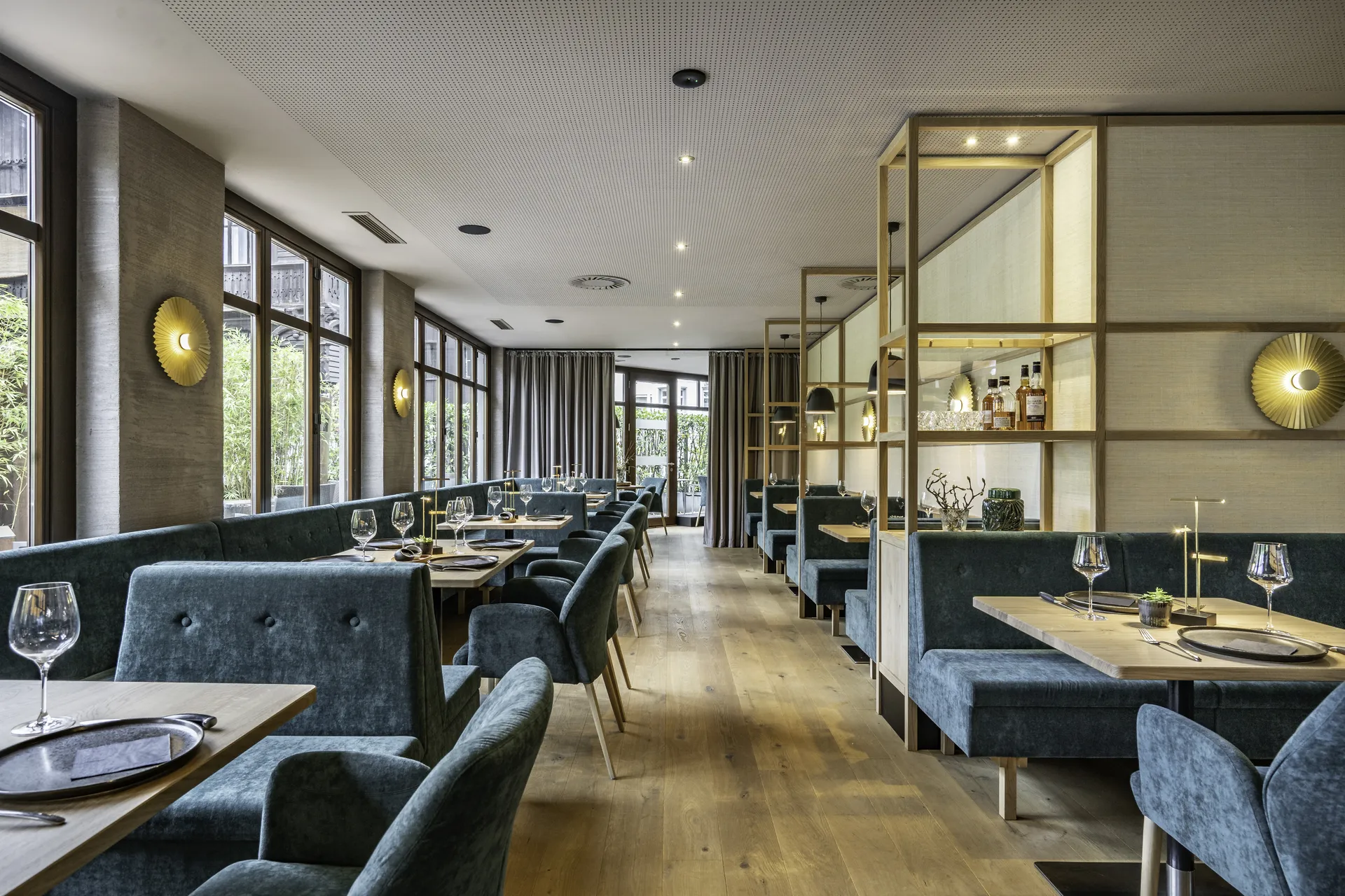 Gastraum des Steakhaus 21 in Dornbirn mit blauen Samtbänken und Sesseln, gedeckten Holztischen, großer Fensterfront und goldenen Wandleuchten.