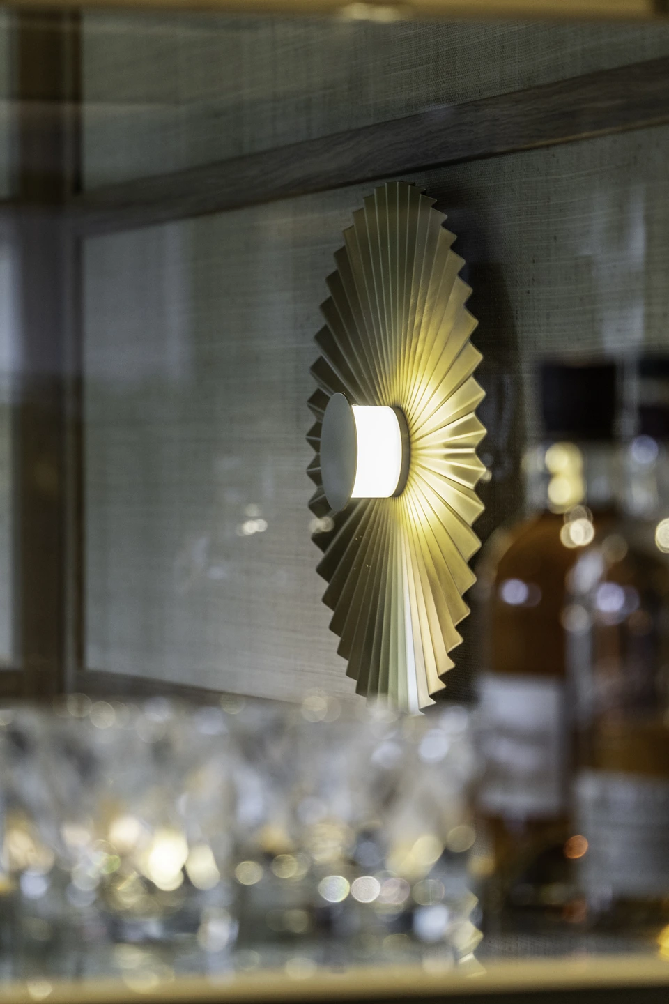 Detailaufnahme einer goldenen Fächer-Wandleuchte im Steakhaus 21 Dornbirn, mit unscharfen Weingläsern im Vordergrund und einer Whiskyflasche im Hintergrund.