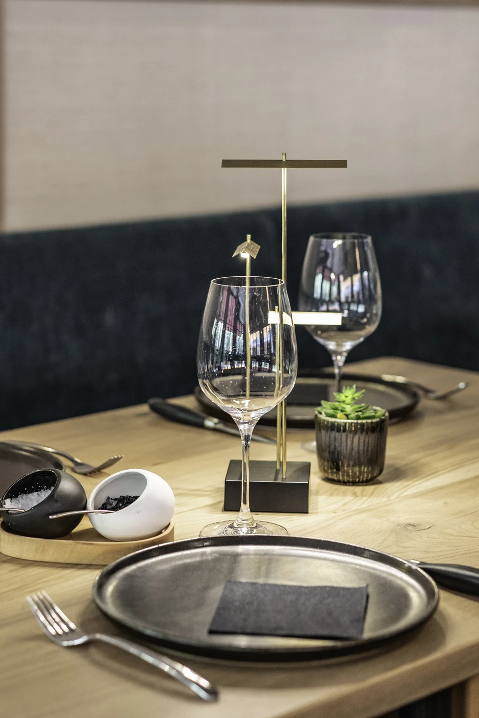 Detailaufnahme eines gedeckten Tisches im Steakhaus 21 Dornbirn mit Weingläsern, schwarzen Tellern, goldener Tischlampe sowie Salz- und Pfefferstreuer auf Holztisch.