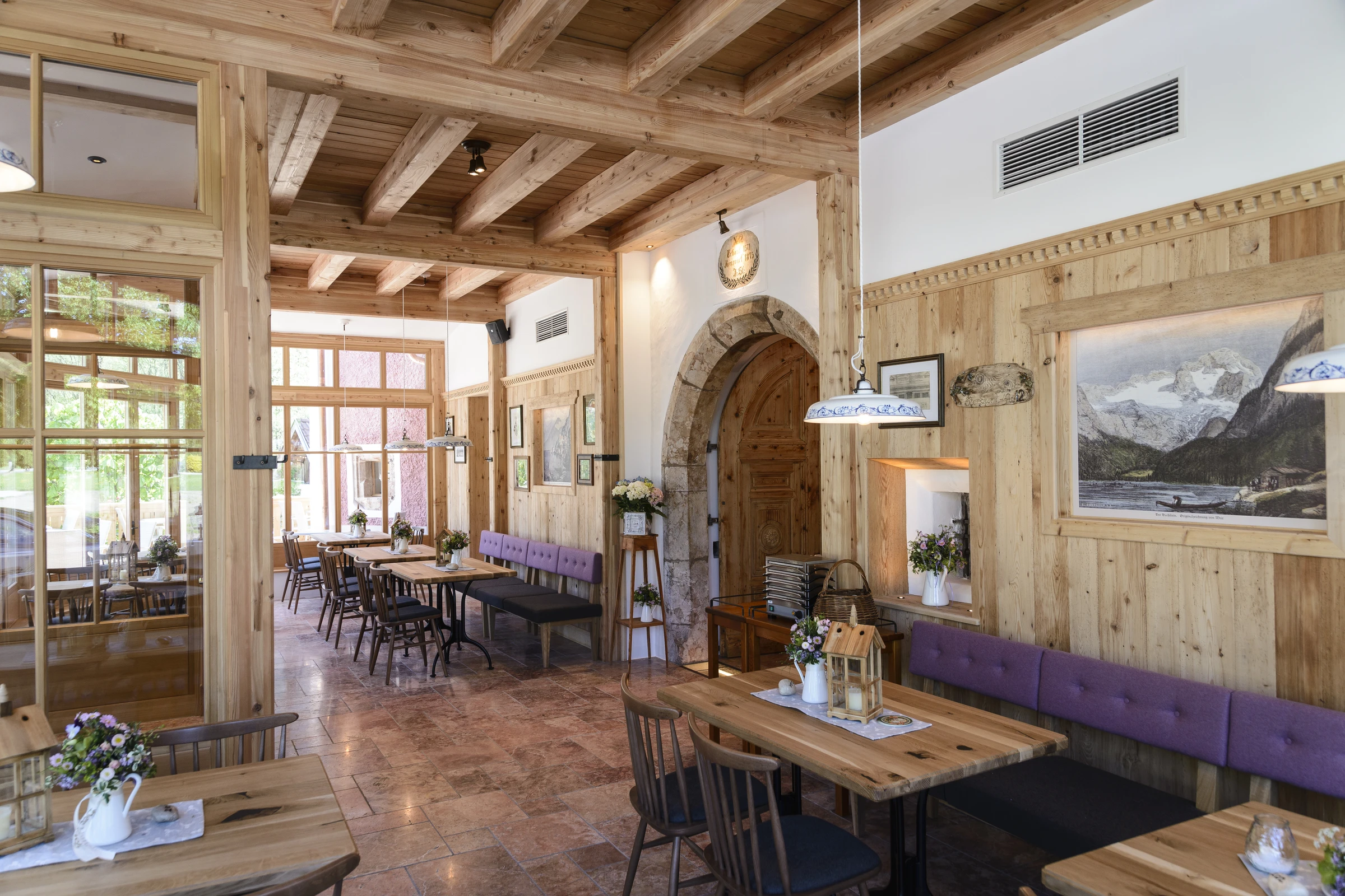 Gastraum des Restaurant Steegwirt mit Holzbalkendecke, Steinrundbogen, lila Sitzbänken, Holztischen und Berggemälden an der Holzvertäfelung.