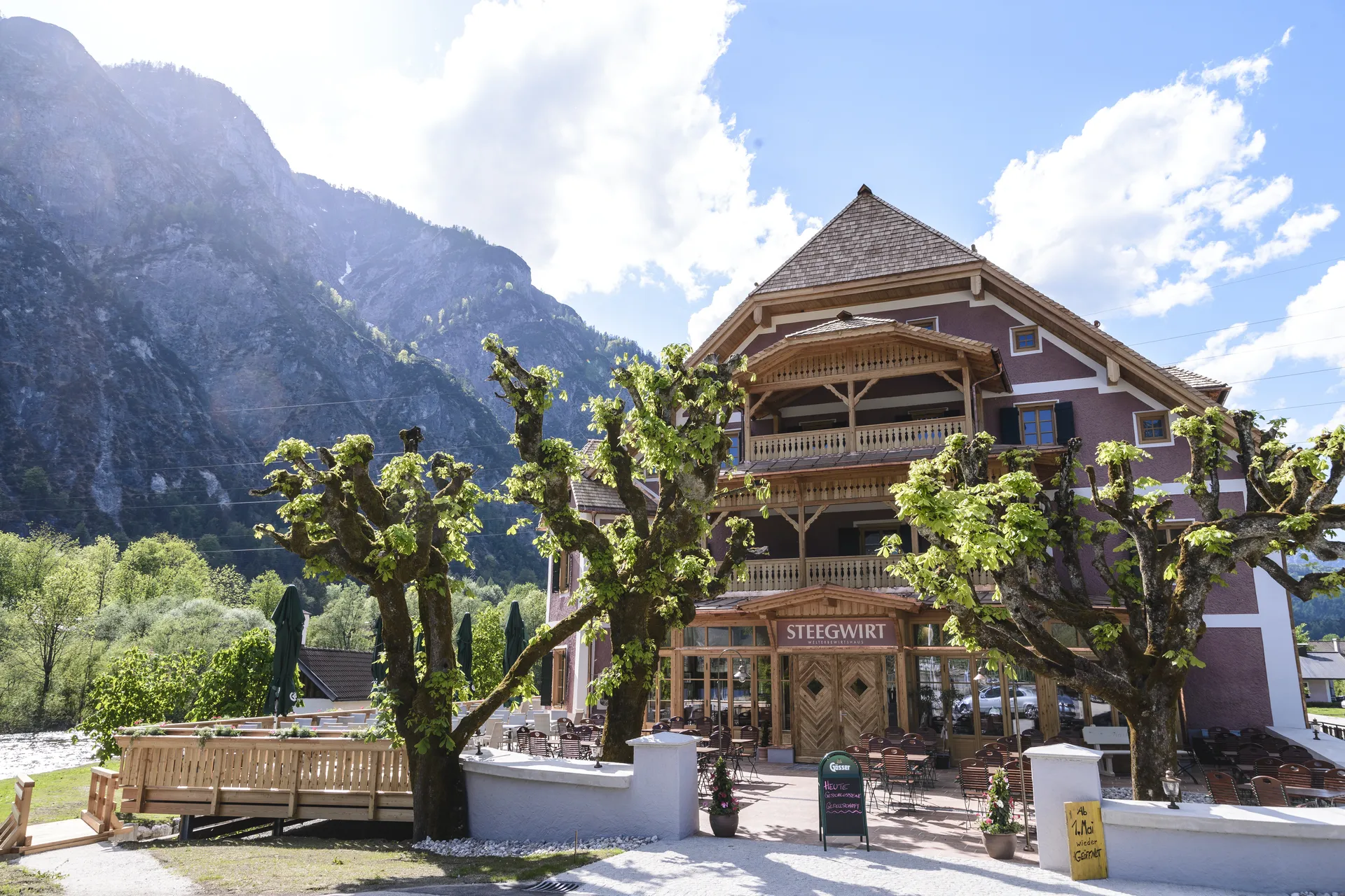 Außenansicht des Steegwirt in Bad Goisern mit historischer Fassade, Holzbalkonen, alten Kastanienbäumen, Terrasse und imposanter Bergkulisse im Hintergrund.