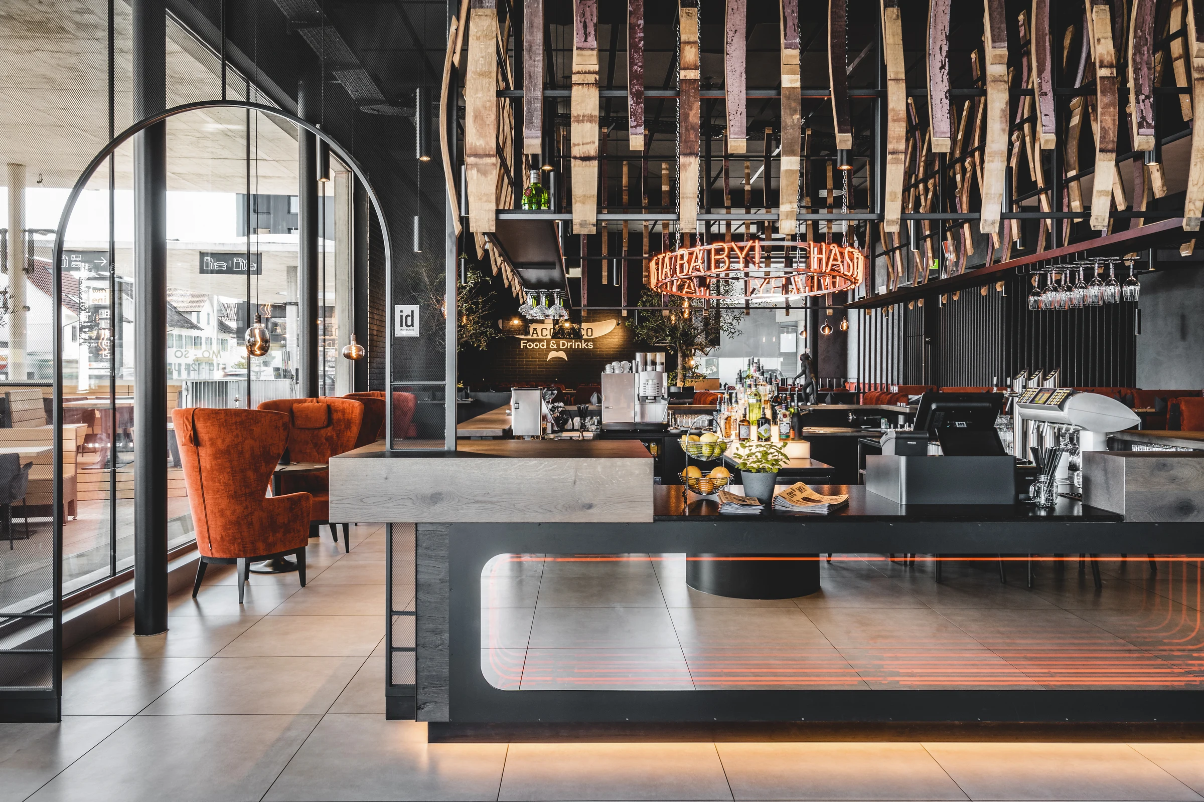 Bar des Taco Taco in Lustenau mit schwarzem Bartresen, orangenem Neonschriftzug, Fassdauben-Deckeninstallation und roten Samtsesseln im Lounge-Bereich.