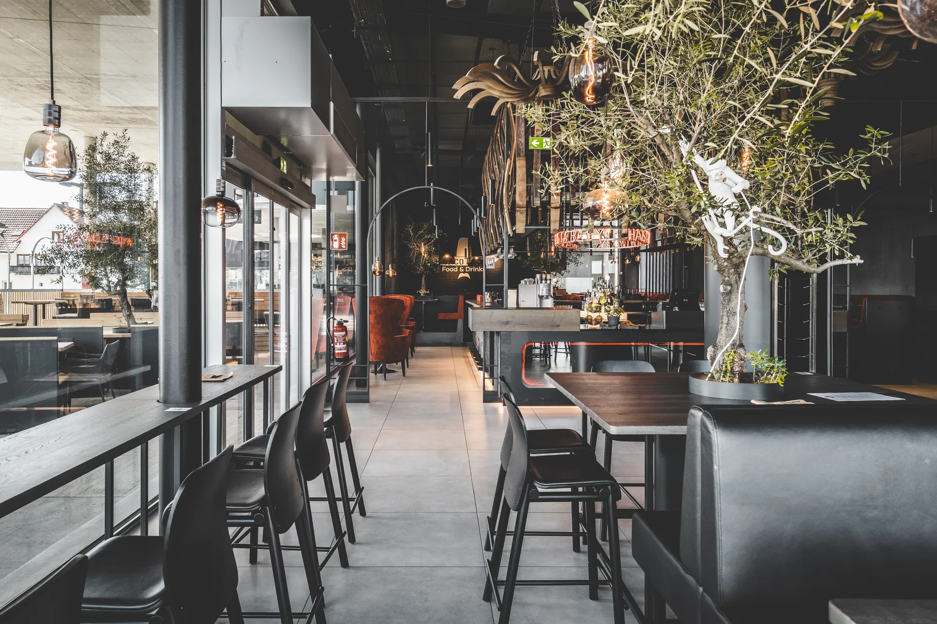 Gesamtansicht des Taco Taco in Lustenau mit Stehtischen, Barhockern, zwei Olivenbäumen, orangem Neonschriftzug und Blick zur Bar im Hintergrund.