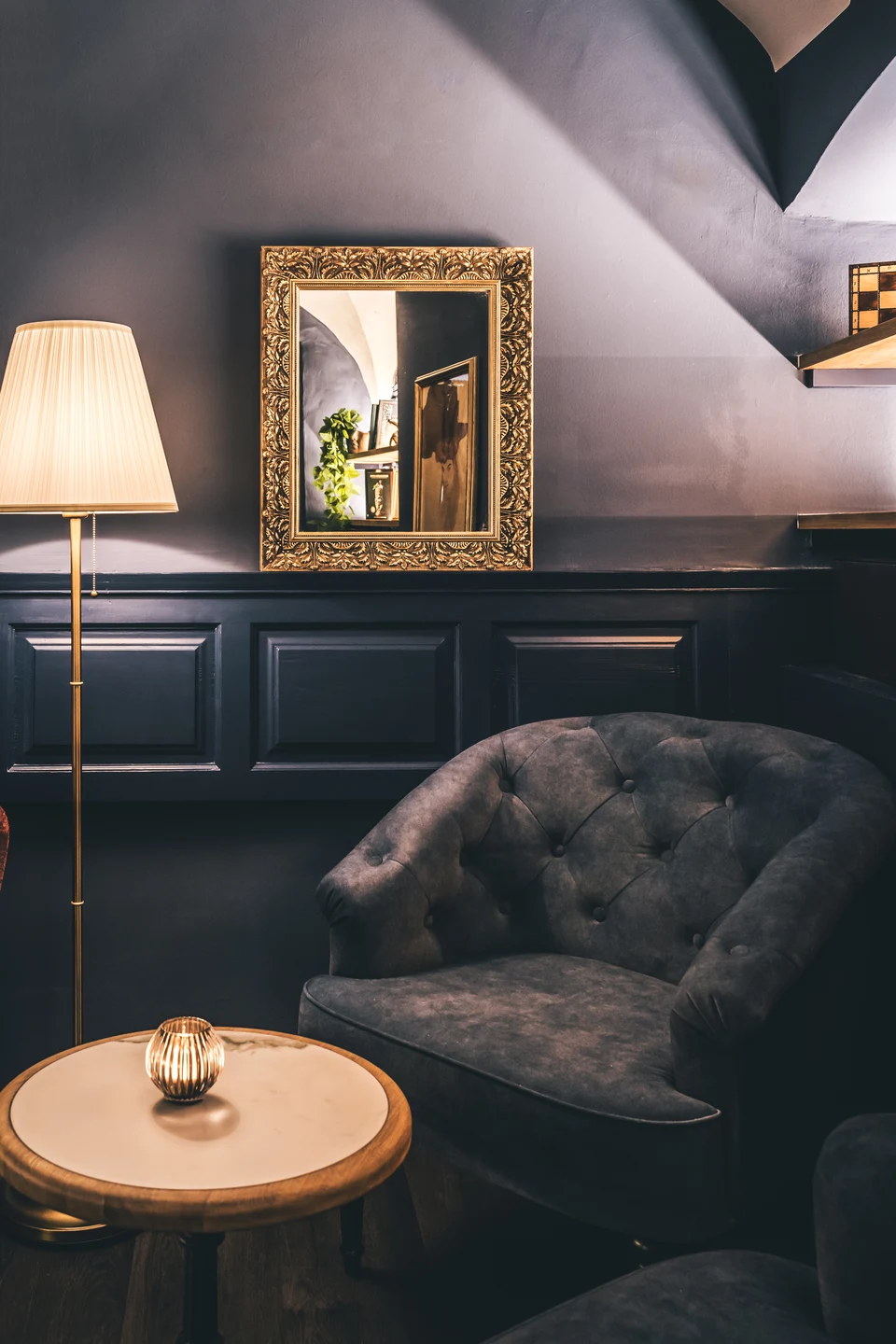Lounge-Bereich der Bar To The Moon in Linz mit grauem Chesterfield-Sessel, goldenem Wandspiegel, Stehlampe und rundem Marmortisch mit Windlicht.