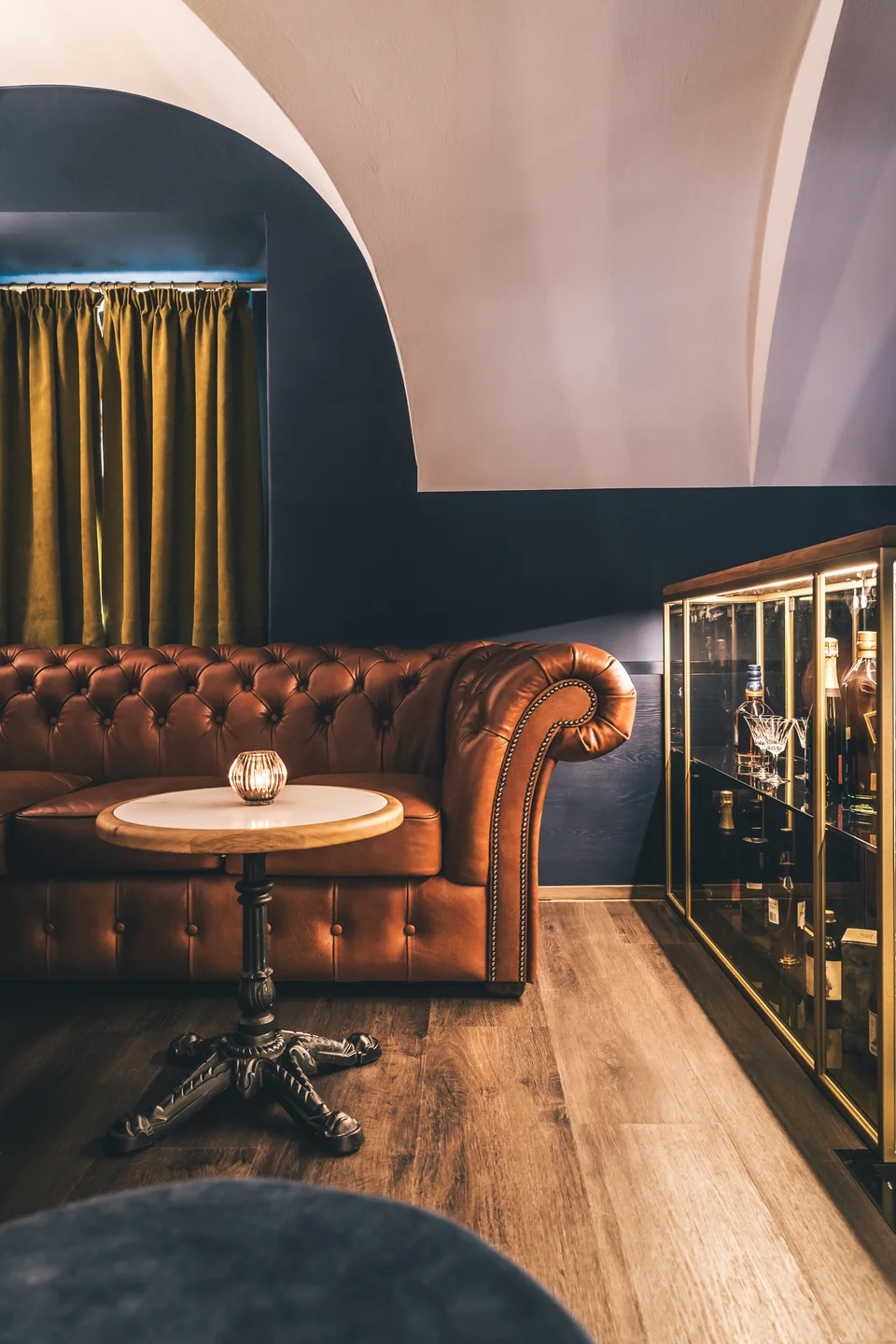 Lounge-Bereich der Bar To The Moon in Linz mit cognacfarbenem Chesterfield-Ledersofa, beleuchtetem Whiskyschrank, Gewölbebogen und goldenen Samtvorhängen.