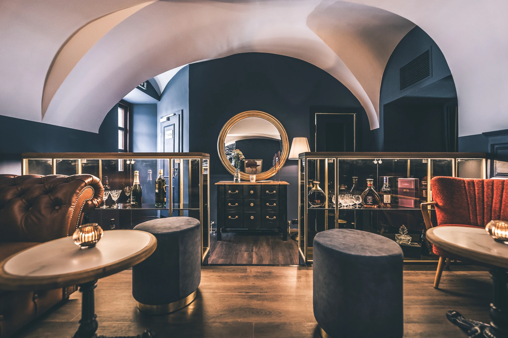 Lounge der Bar To The Moon in Linz mit Kreuzgewölbe, zwei beleuchteten Whiskyschränken, cognacfarbenem Chesterfield-Sofa, goldenem Rundspiegel und antiker Kommode.