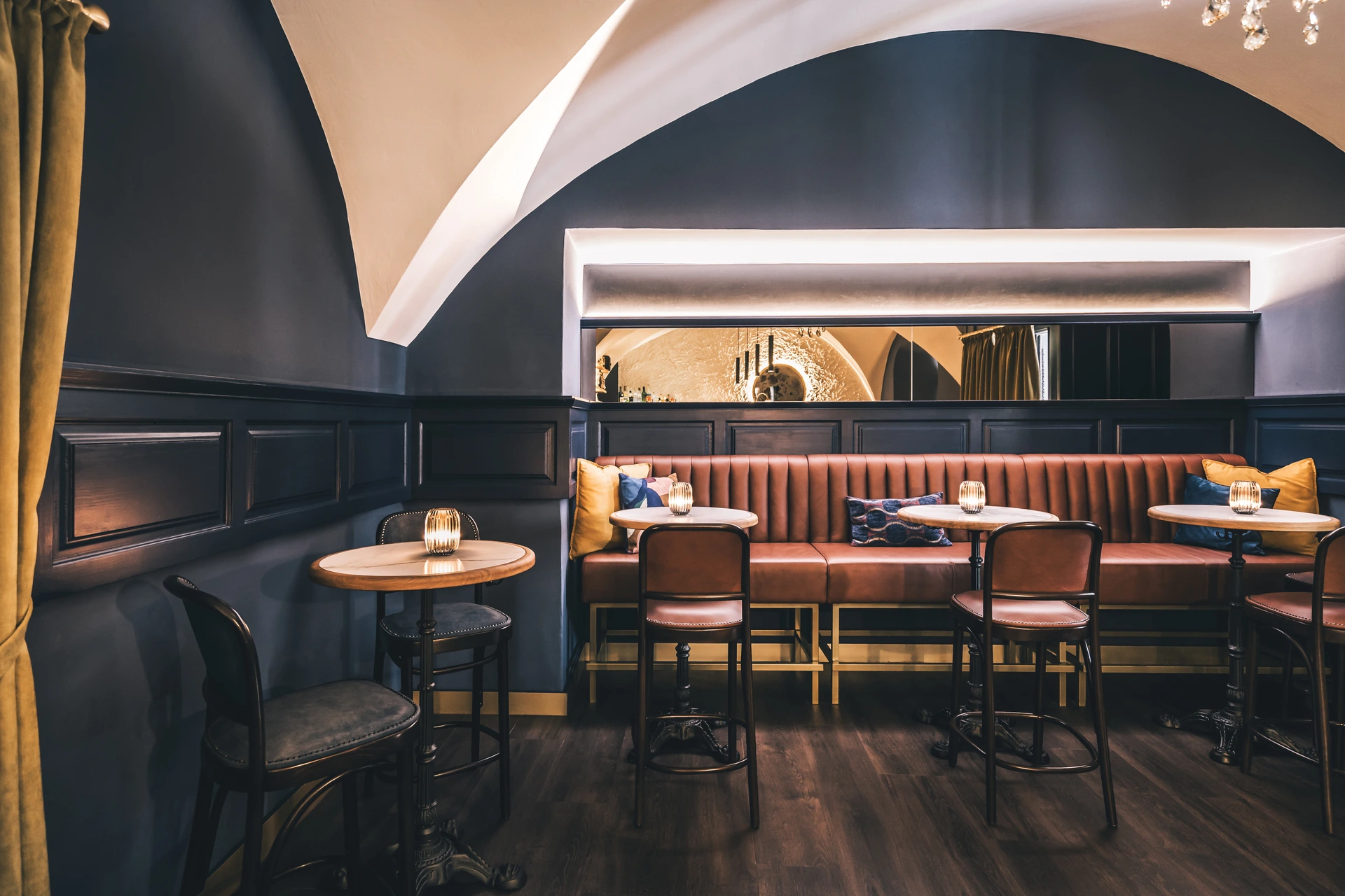 Sitzbereich der Bar To The Moon in Linz mit cognacfarbener Lederbank, bunten Kissen, Marmortischen, Windlichtern, Gewölbebogen und Spiegelblick zur Bar.