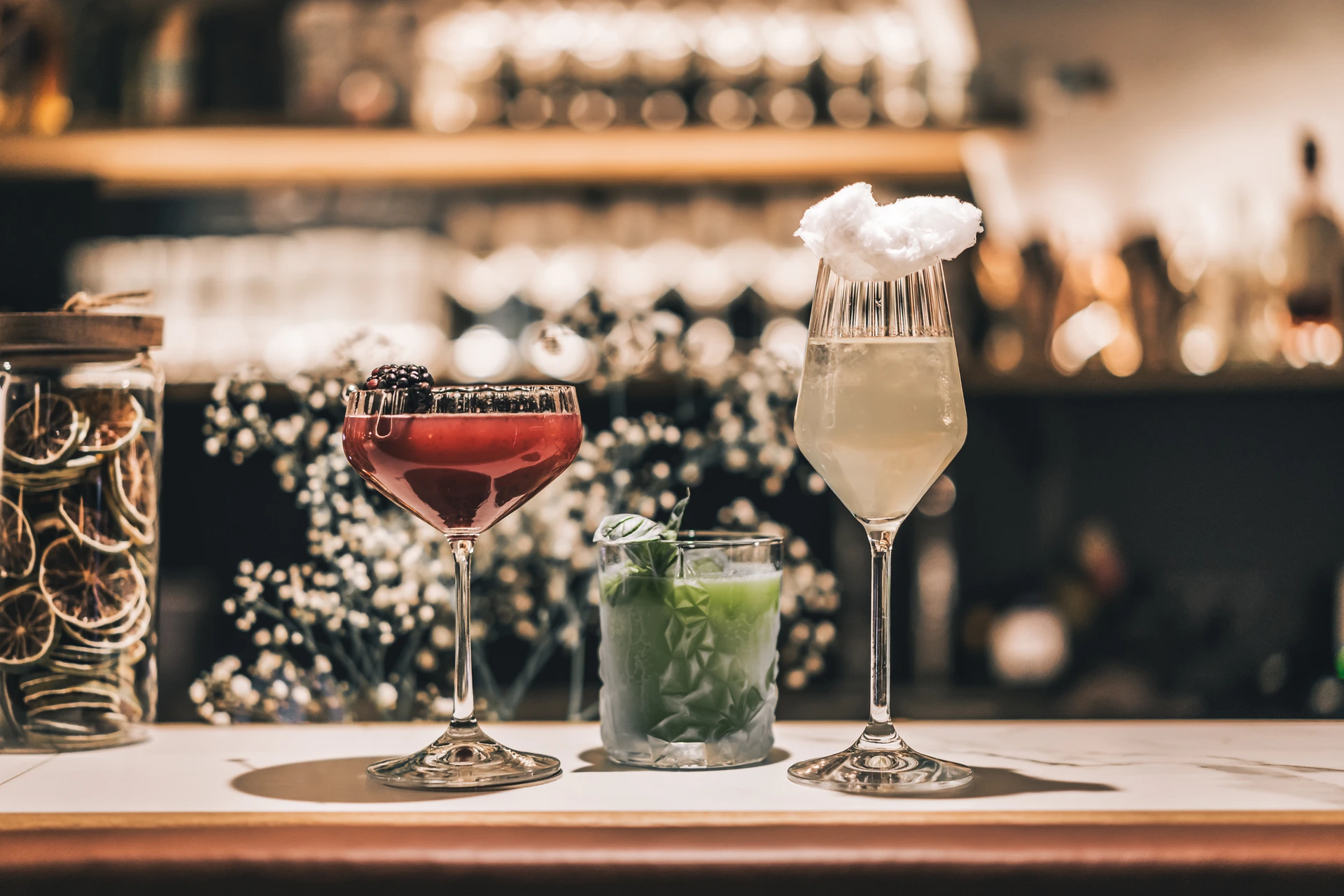 Drei Cocktails auf dem Marmortresen der Bar To The Moon in Linz – ein Brombeer-Coupe, ein grüner Basilikum-Tumbler und ein Sekt-Flute mit Zuckerwatte, dekoriert mit Schleierkraut.