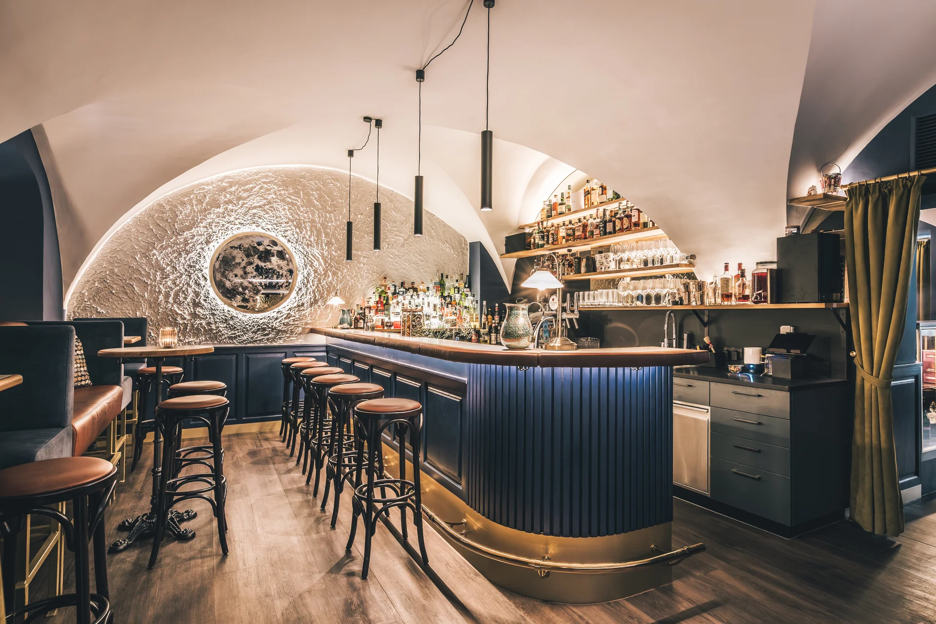 Gesamtansicht der Bar To The Moon in Linz mit blauem Rippentresen, beleuchtetem Mondmotiv-Wandrelief, großem Spirituosenregal, braunen Barhockern und Gewölbedecke.