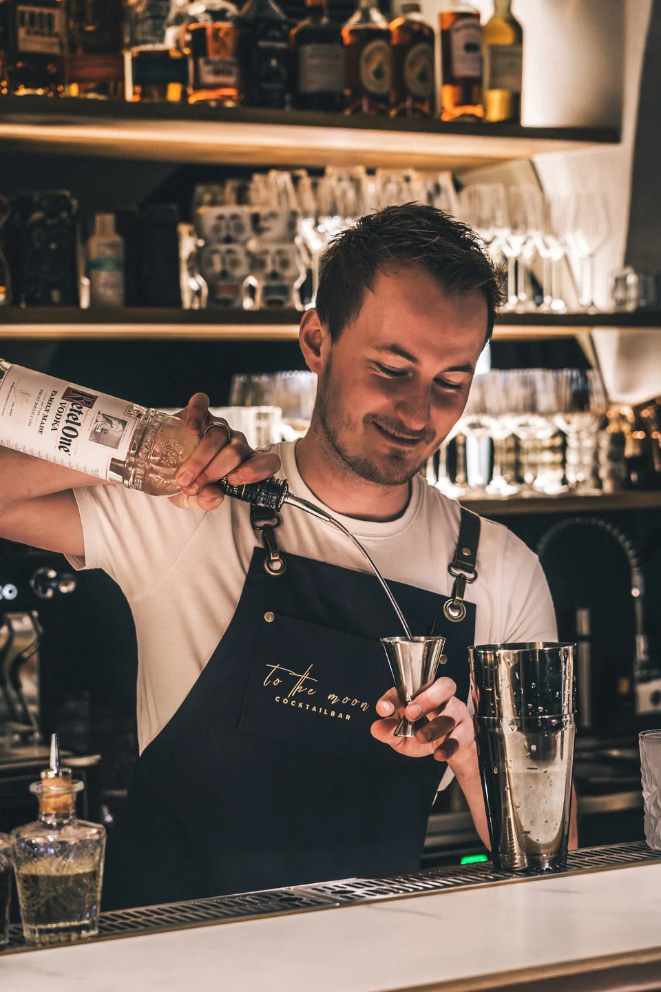 Barkeeper der To The Moon Cocktailbar in Linz schüttet Ketel One Vodka in einen Jigger, trägt schwarze Schürze mit Logo vor beleuchtetem Spirituosenregal.