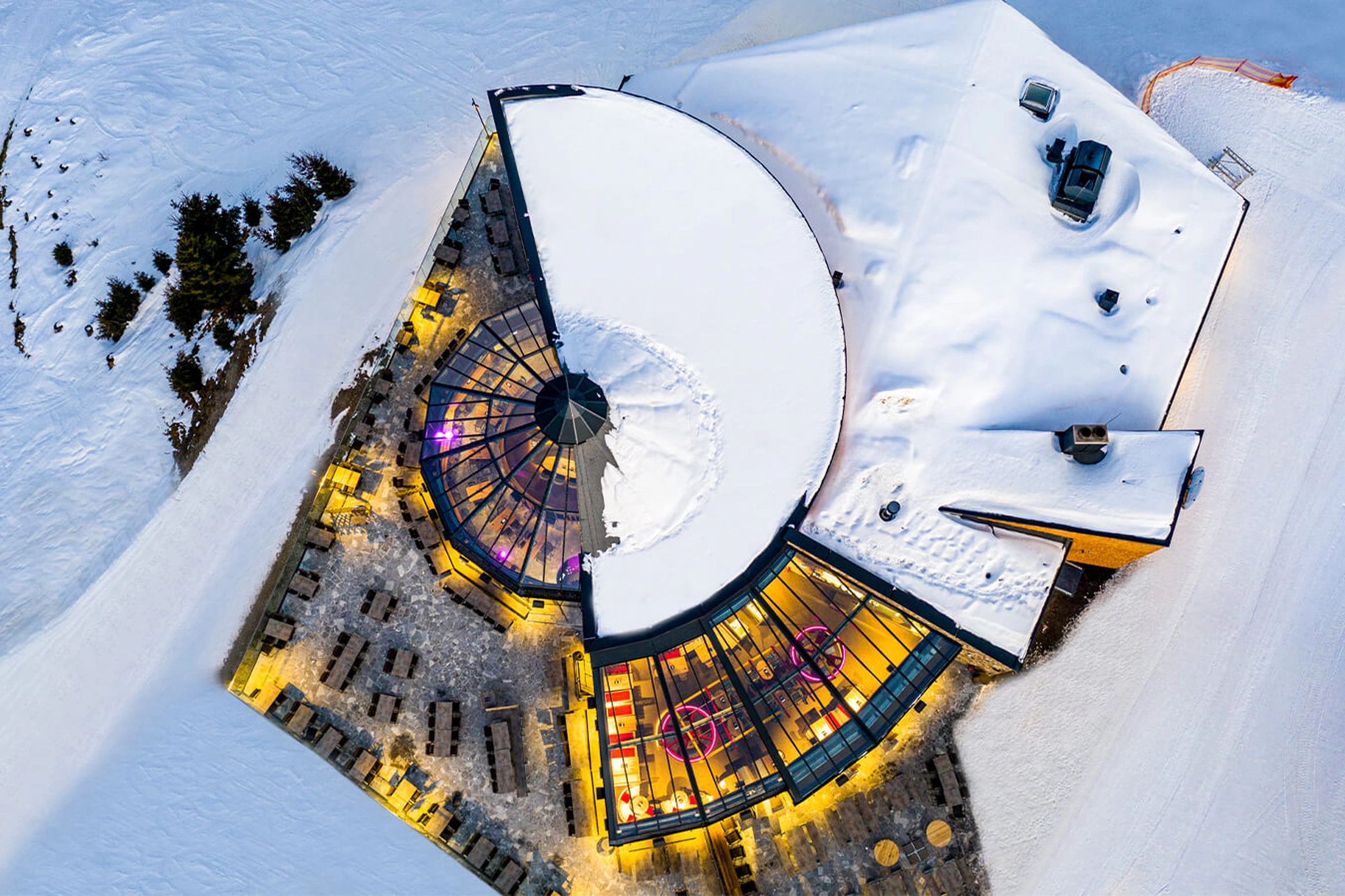 Drohnenaufnahme der Umbrella Bar in Gerlos aus der Vogelperspektive – rundes Gebäude mit teilweise verschneitem Glasdach und warmem Innenbeleuchtung, umgeben von verschneiter Winterlandschaft und Außenterrasse.