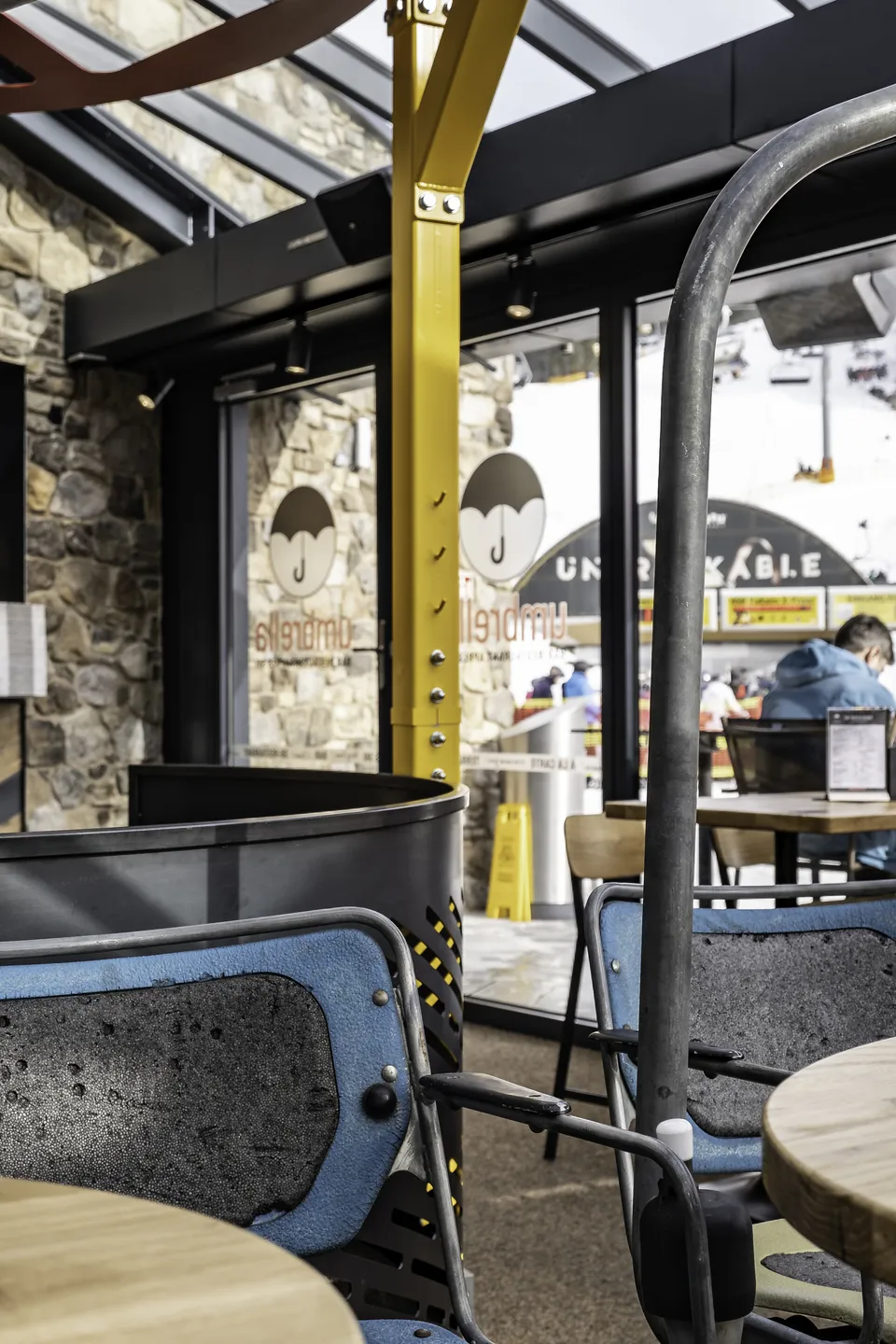 Innenraum der Umbrella Bar in Gerlos mit repurposed Sessellift-Sitzen als Stühle, Holztischen, gelben Stahlträgern und Glasfront mit Blick auf die verschneite Skipiste.