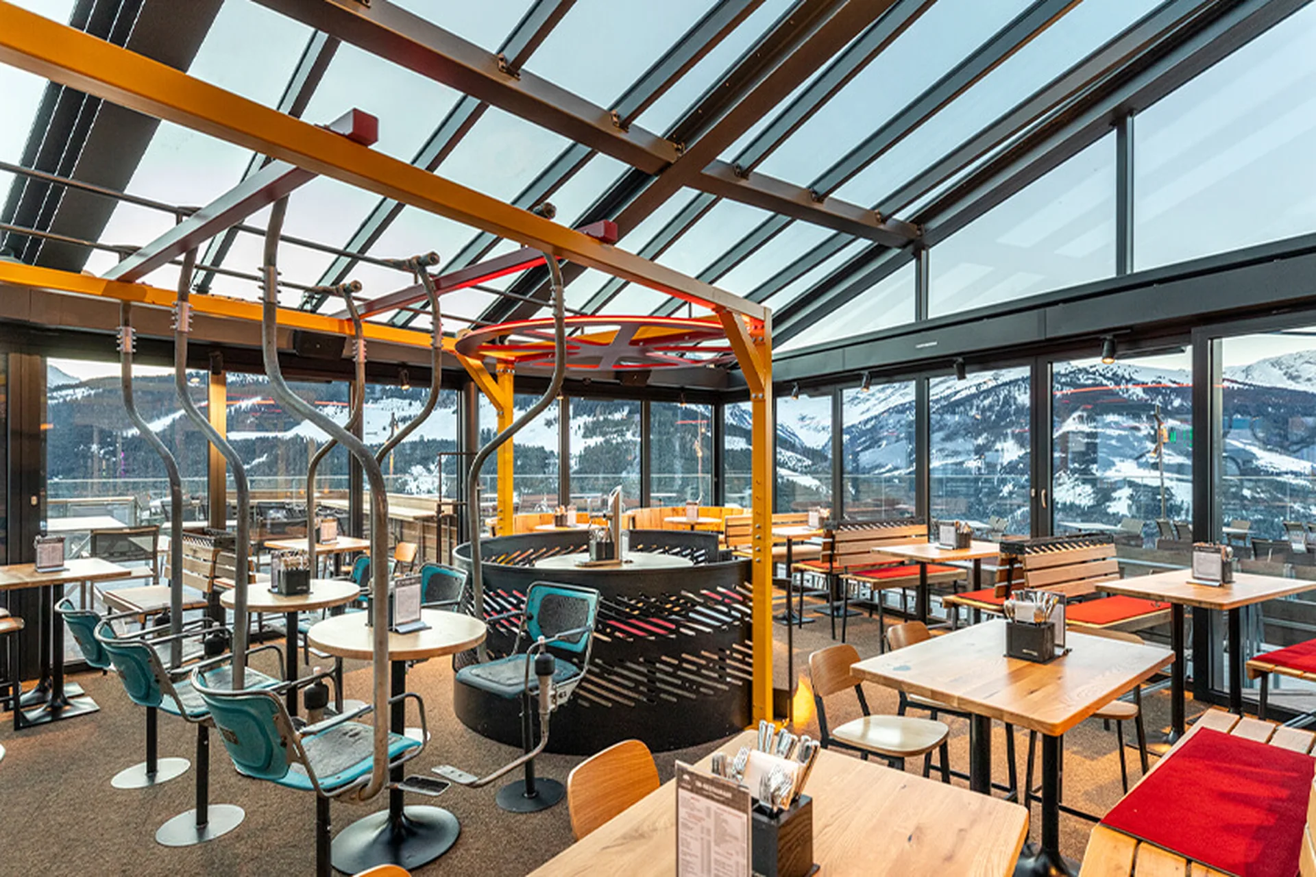 Gastraum der Umbrella Bar in Gerlos mit großer Glasfront, Bergpanorama und Skipiste, Sessellift-Sitzen, roten Sitzkissen und gelben Stahlträgern unter dem Glasdach.