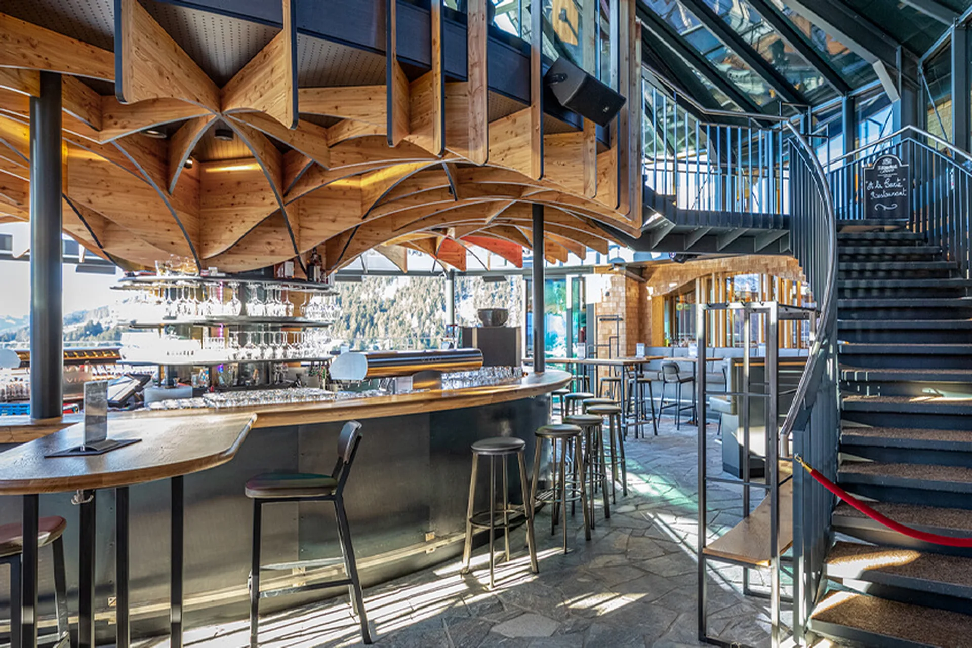 Innenraum der Umbrella Bar in Gerlos mit geschwungenem Bartresen, strukturiertem Holzrippendach, schwarzen Barhockern, Wendeltreppe und Panoramablick auf die verschneite Berglandschaft.