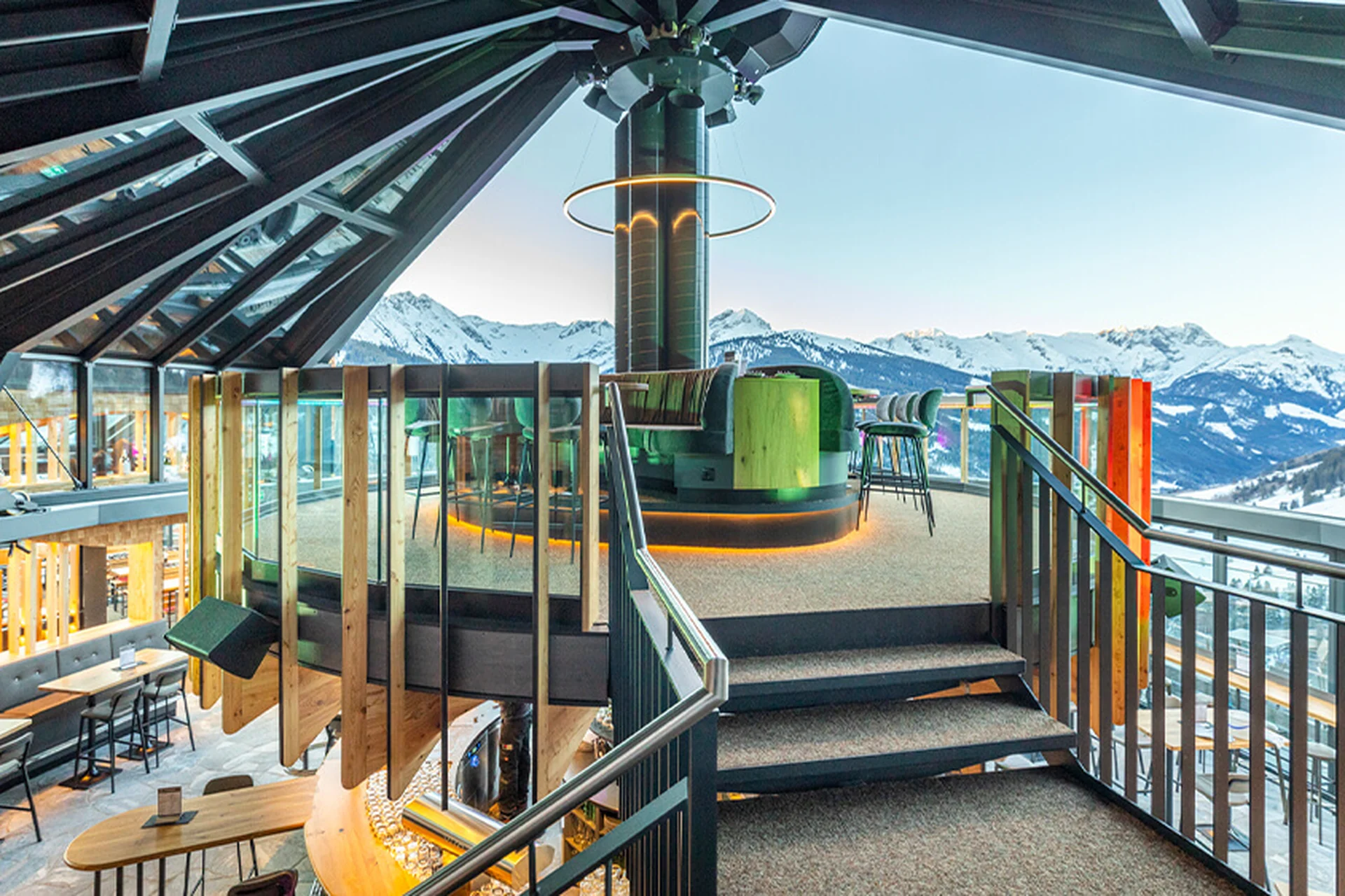 Blick vom Obergeschoss der Umbrella Bar in Gerlos auf die runde Bar mit grünen Barhockern, hängender Ringleuchte und Treppenaufgang, im Hintergrund Panoramablick auf die verschneite Zillertaler Bergkulisse.