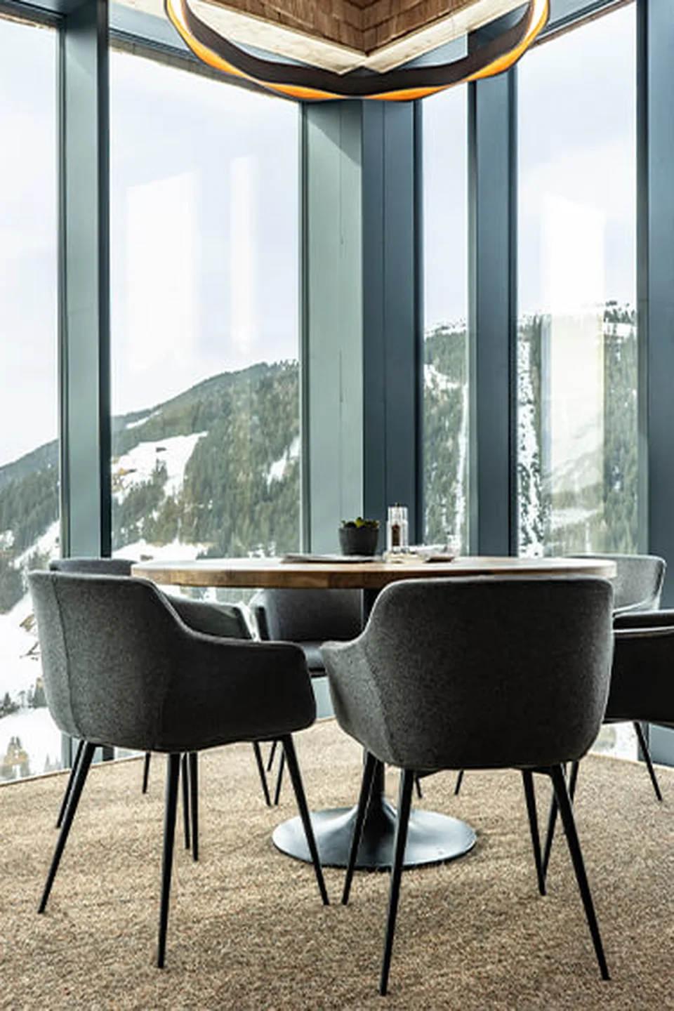 Sitzbereich der Umbrella Bar in Gerlos mit rundem Holztisch, grauen Polstersesseln, bodentiefen Panoramafenstern mit Blick auf die verschneite Berglandschaft und hängender Ringleuchte.