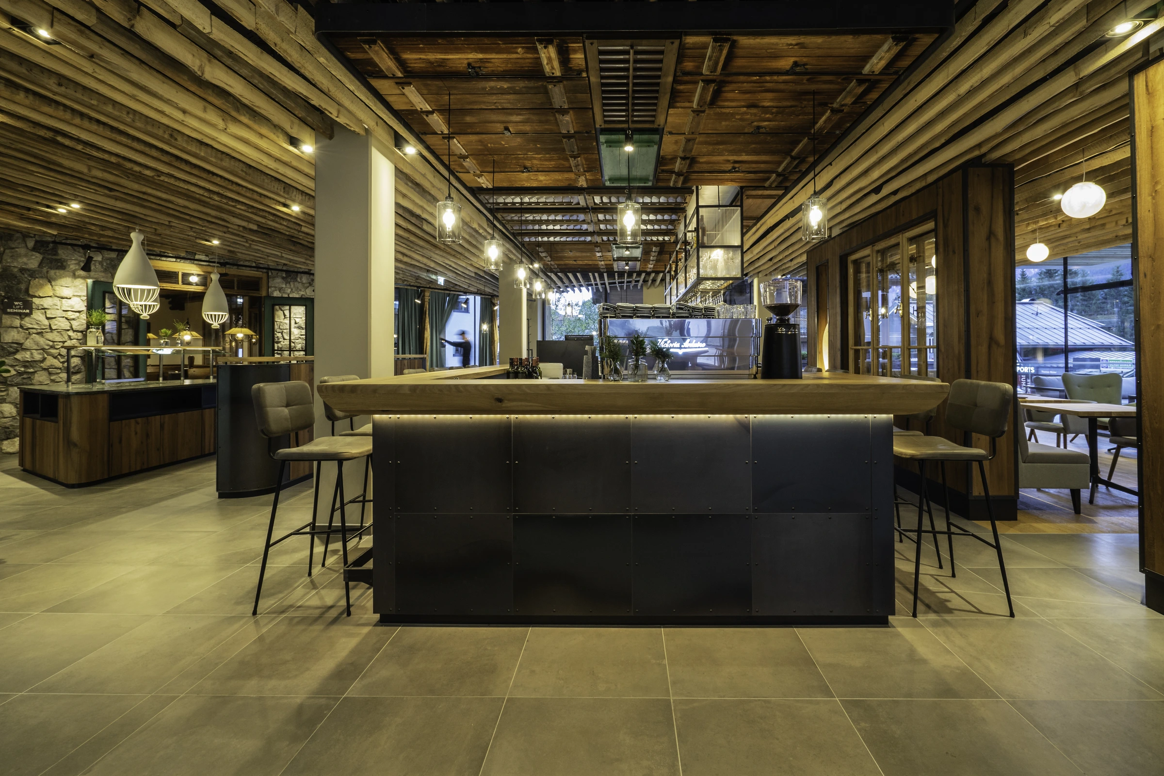Bar der Venediger Lodge in Neunkirchen mit zentralem Naturholztresen, Stahlverkleidung mit indirekter Beleuchtung, rustikaler Altholzdecke, Victoria-Arduino-Espressomaschine und hängenden Glaslampen.