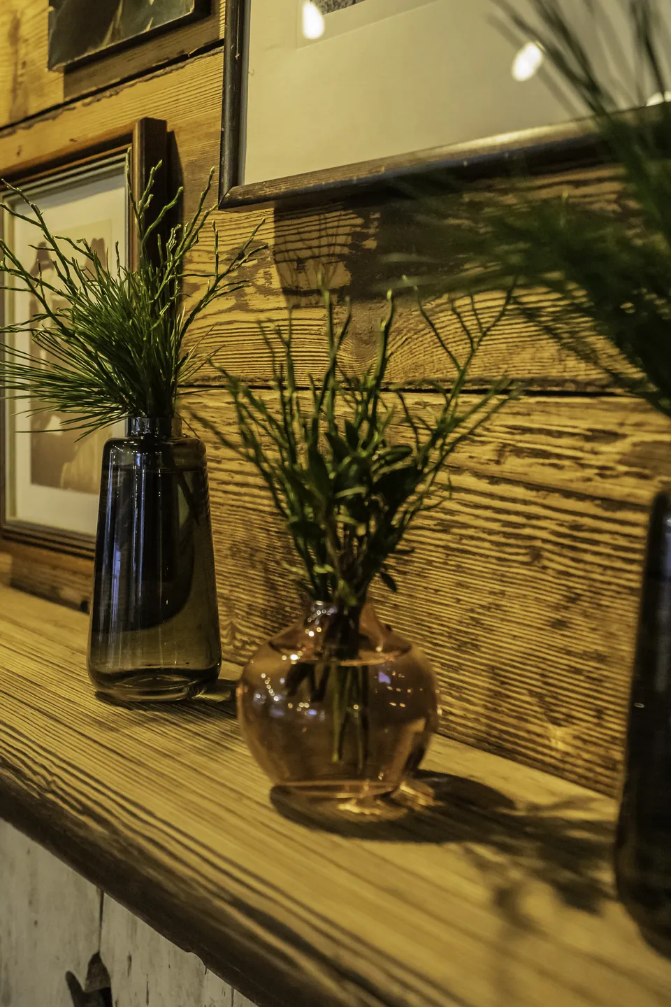 Detailaufnahme dreier Glasvasen mit Kräuterdeko auf einem Naturholzregal vor rustikaler Altholzwand mit vintage Bilderrahmen in der Venediger Lodge Neunkirchen.