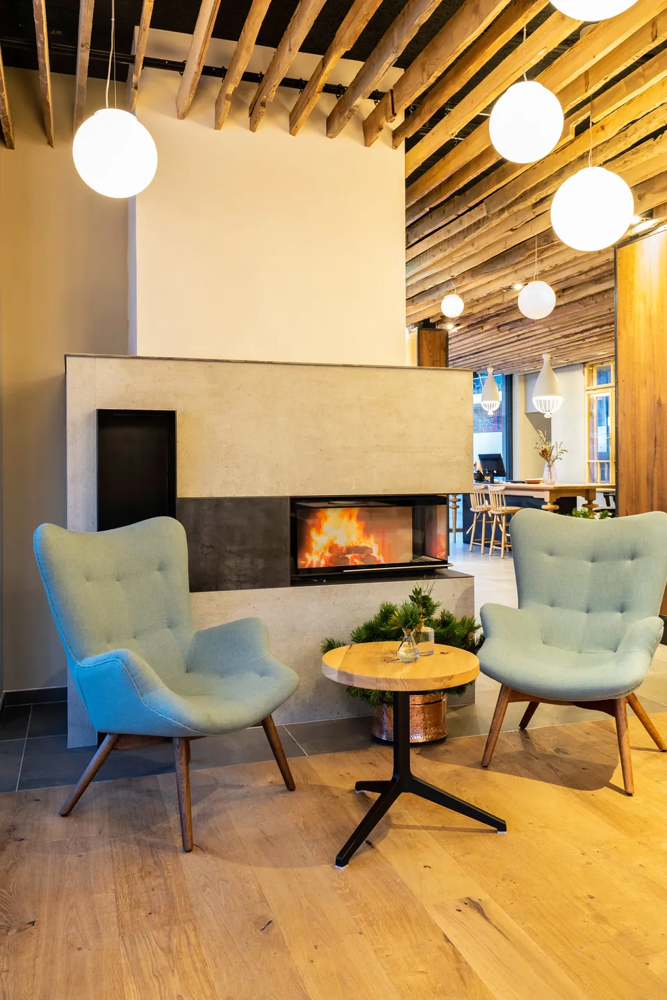 Loungebereich der Venediger Lodge Neunkirchen mit zwei mintgrünen Ohrensesseln, modernem Kamin mit offenem Feuer, rundem Holzbeistelltisch, Tannendeko und hängenden Kugelleuchten vor rustikaler Altholzbalkendecke.