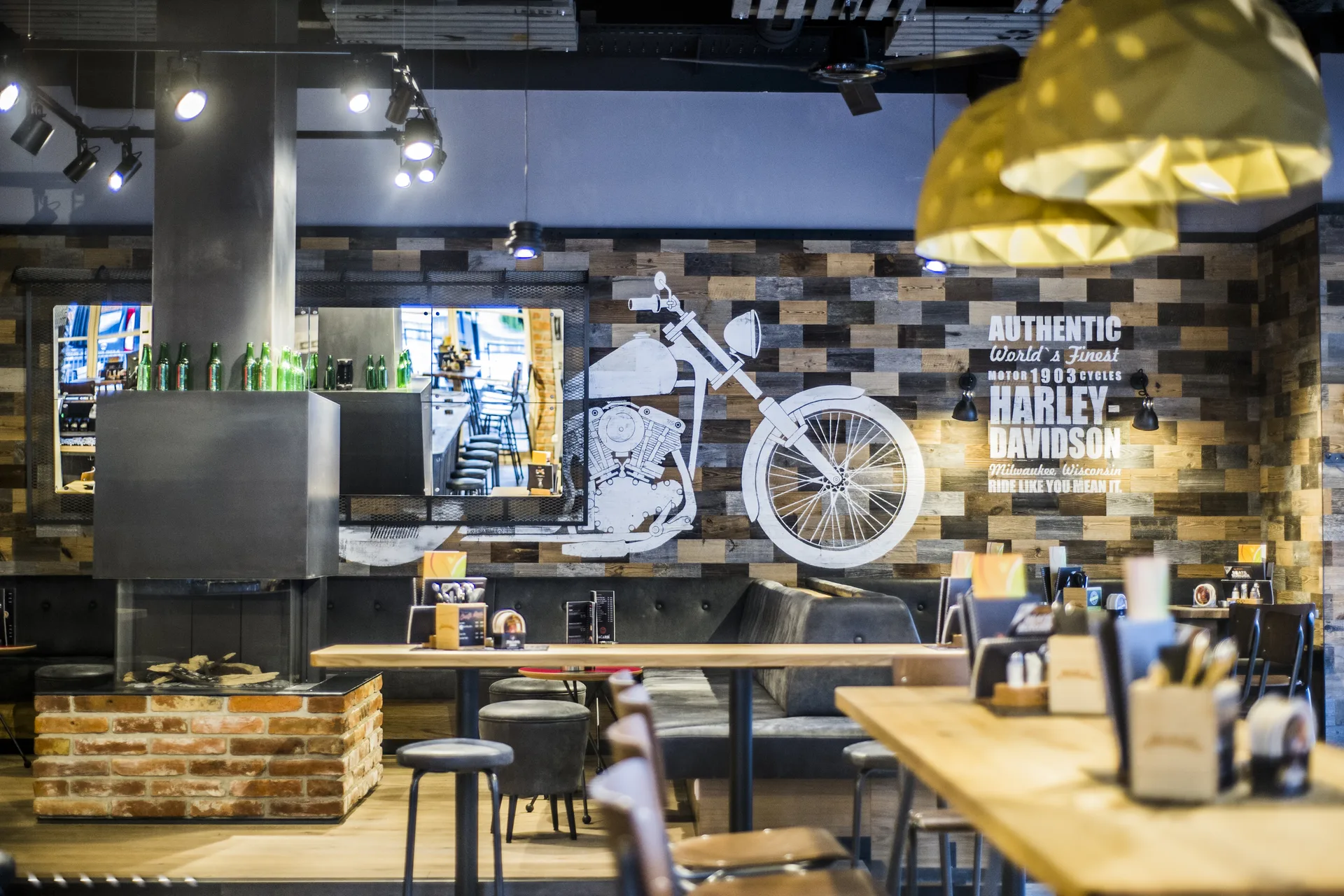 Gastraum der Werkstatt Kufstein mit großem Harley-Davidson-Wandbild auf rustikaler Altholzwand, gedeckten Holztischen, grauen Polstersesseln, Backsteinpodest und goldener Origami-Pendelleuchte im Vordergrund.