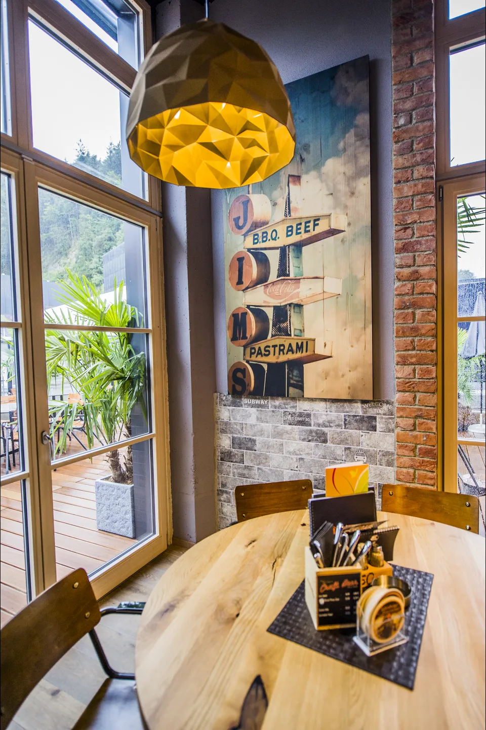 Sitzbereich der Werkstatt Kufstein mit rundem Naturholztisch, goldener Origami-Pendelleuchte, Wegweiser-Wandbild mit BBQ-Beef- und Pastrami-Schildern, rustikaler Ziegelwand und Blick auf die bepflanzte Terrasse.