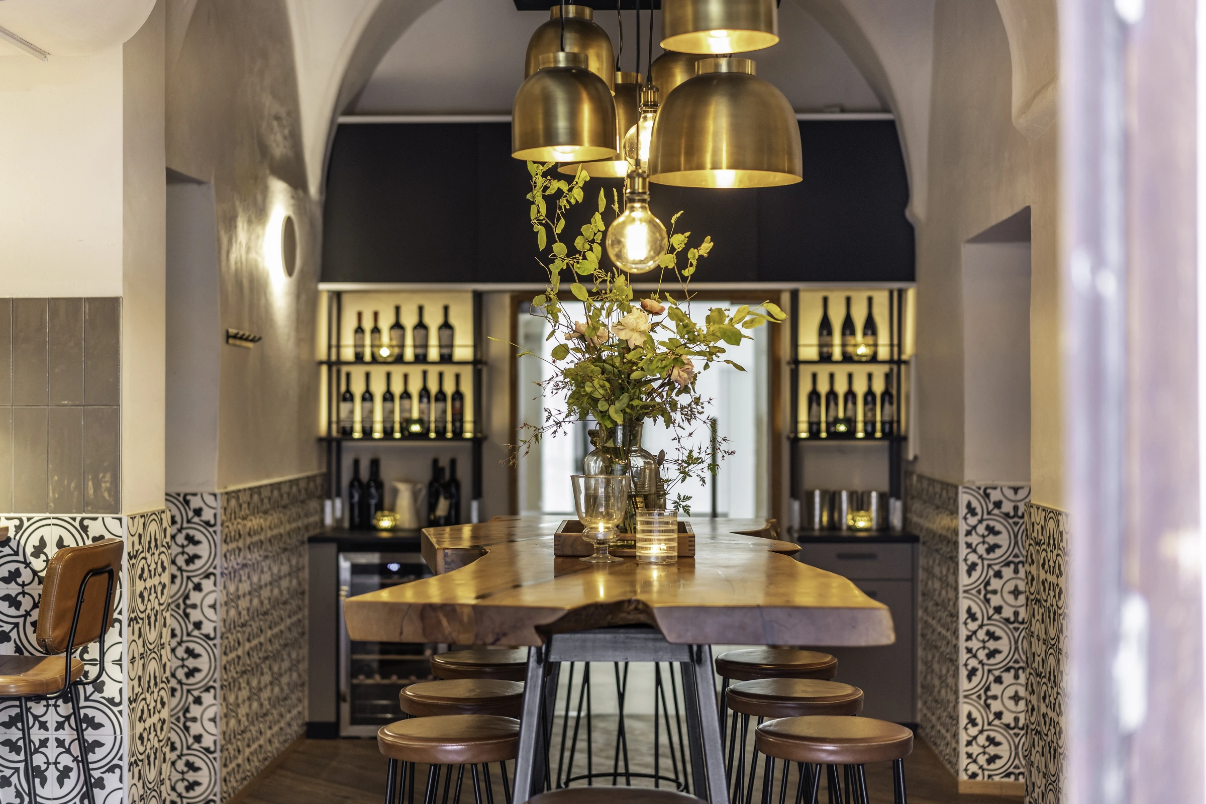 Weinbar des Wirt am Platzl in Garsten mit langem Naturholztisch, Barhockern, goldenen Messing-Pendelleuchten, beleuchtetem Weinregal, Blumengesteck und marokkanischen Wandfliesen unter historischem Gewölbebogen.