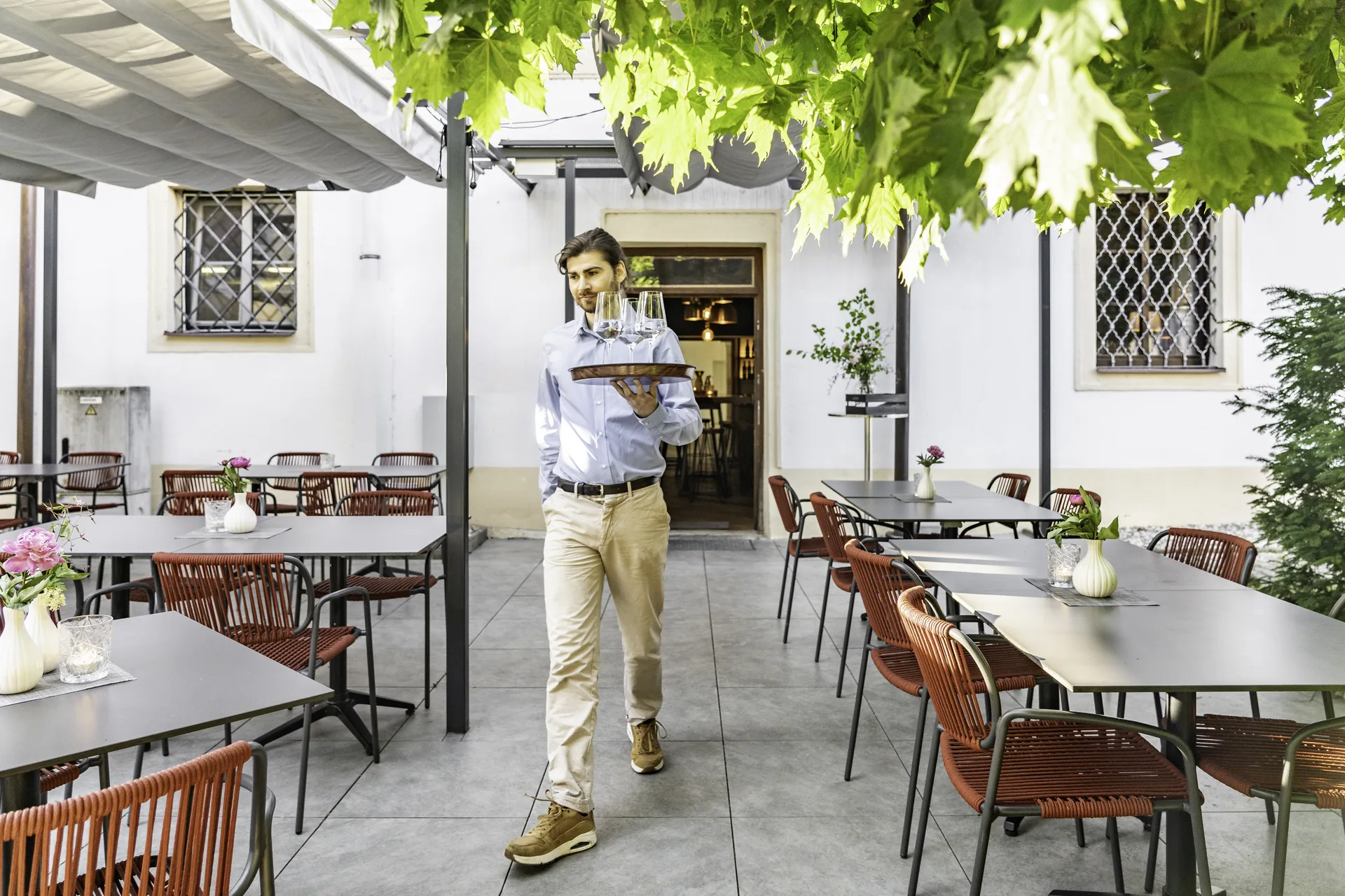 Terrasse des Wirt am Platzl Garsten mit Kellner beim Servieren von Weingläsern auf Holztablett, roten Flechtsessel, dunklen Tischen, Pergola mit Weinblättern und historischer Klosterfassade im Hintergrund.